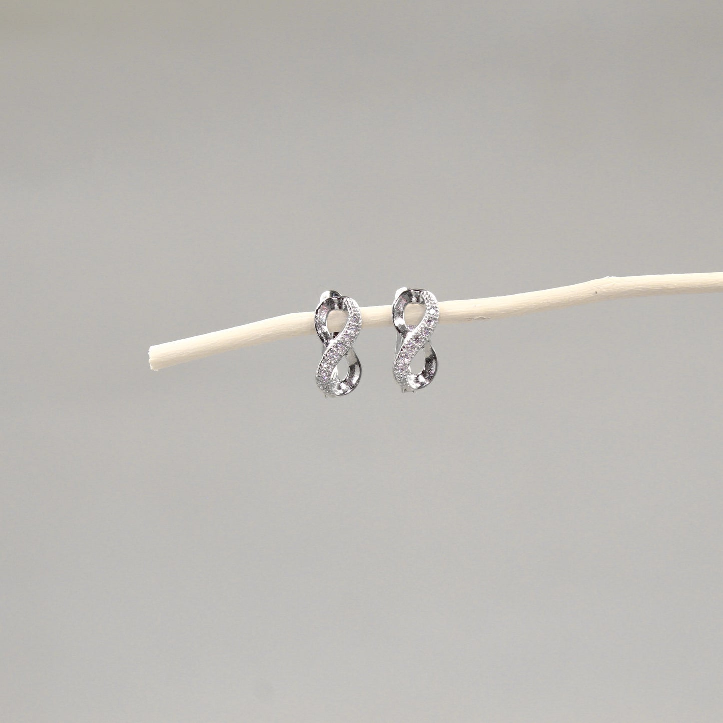 Dos pendientes de plata en forma de lazo infinito con incrustaciones de cristales, colocados delicadamente sobre una varilla de madera pálida, con un fondo gris claro que realza su diseño brillante y elegante.