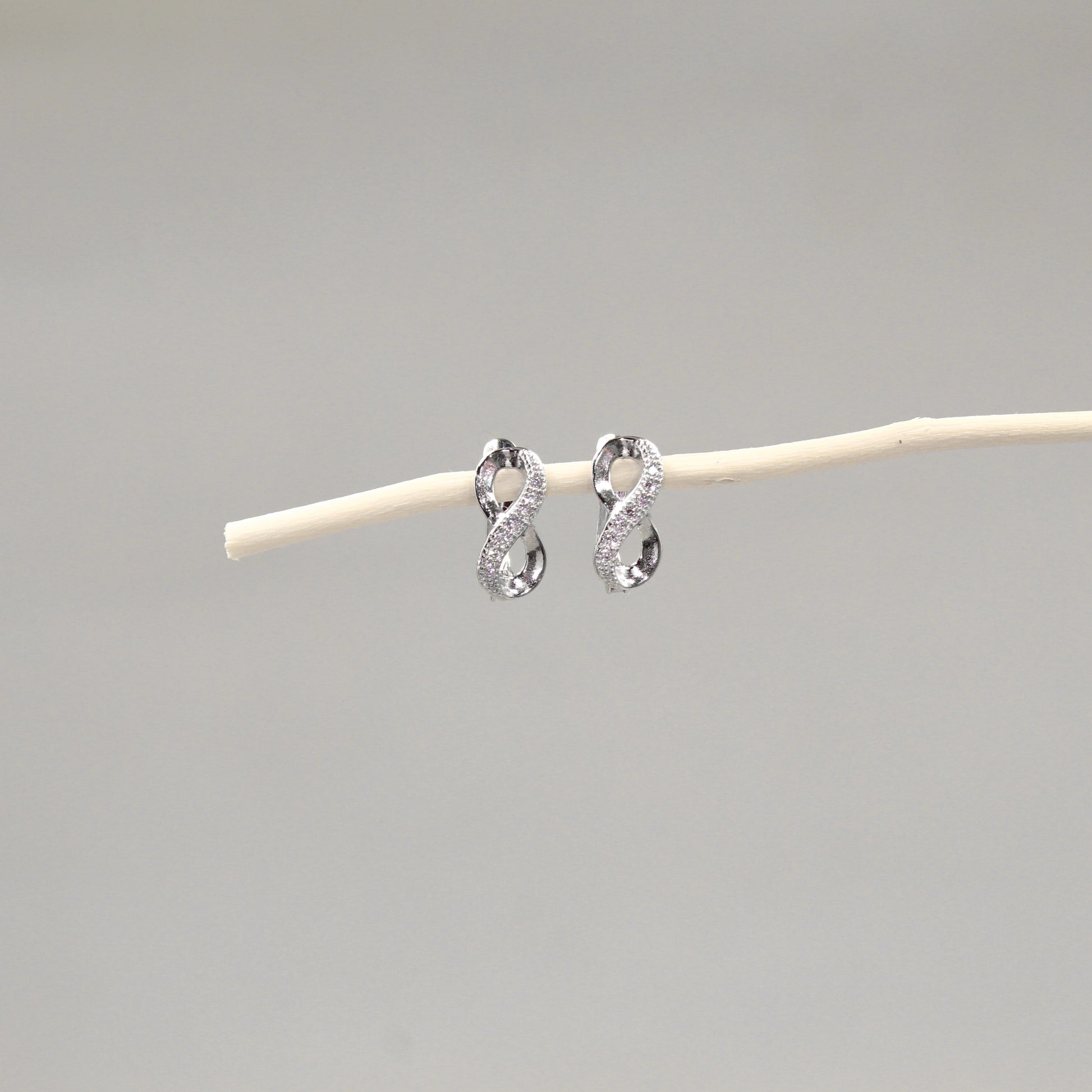 Dos pendientes de plata en forma de lazo infinito con incrustaciones de cristales, colocados delicadamente sobre una varilla de madera pálida, con un fondo gris claro que realza su diseño brillante y elegante.