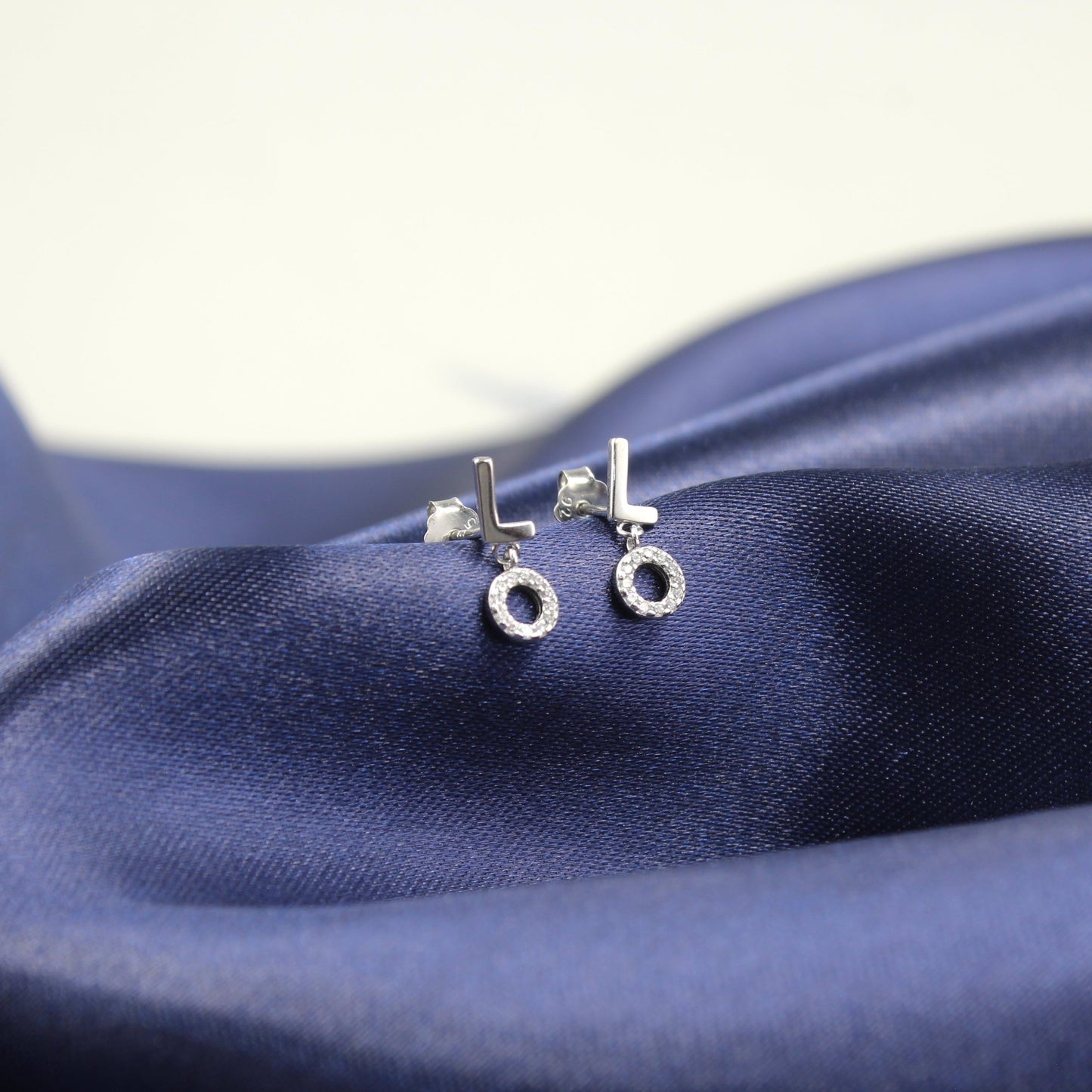 par de aretes de plata con diseño moderno, presentando la palabra 'LOVE' dividida entre ambos. Las letras 'L' y 'O' en un arete y las letras 'V' y 'E' en el otro, cada letra acompañada por un pequeño círculo de circonias cúbicas. Los aretes están colocados sobre un fondo de tela azul satinada, resaltando su brillo y diseño contemporáneo.