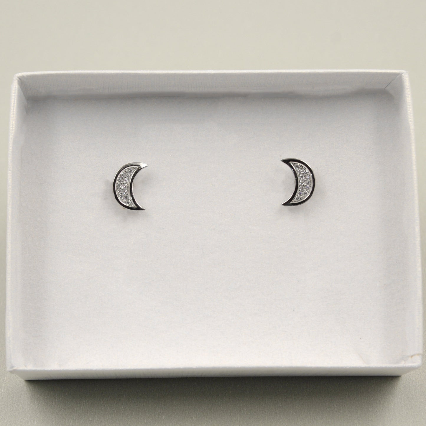 par de aretes de plata con forma de luna creciente, incrustados con pequeños cristales, presentados en una caja blanca con fondo gris.