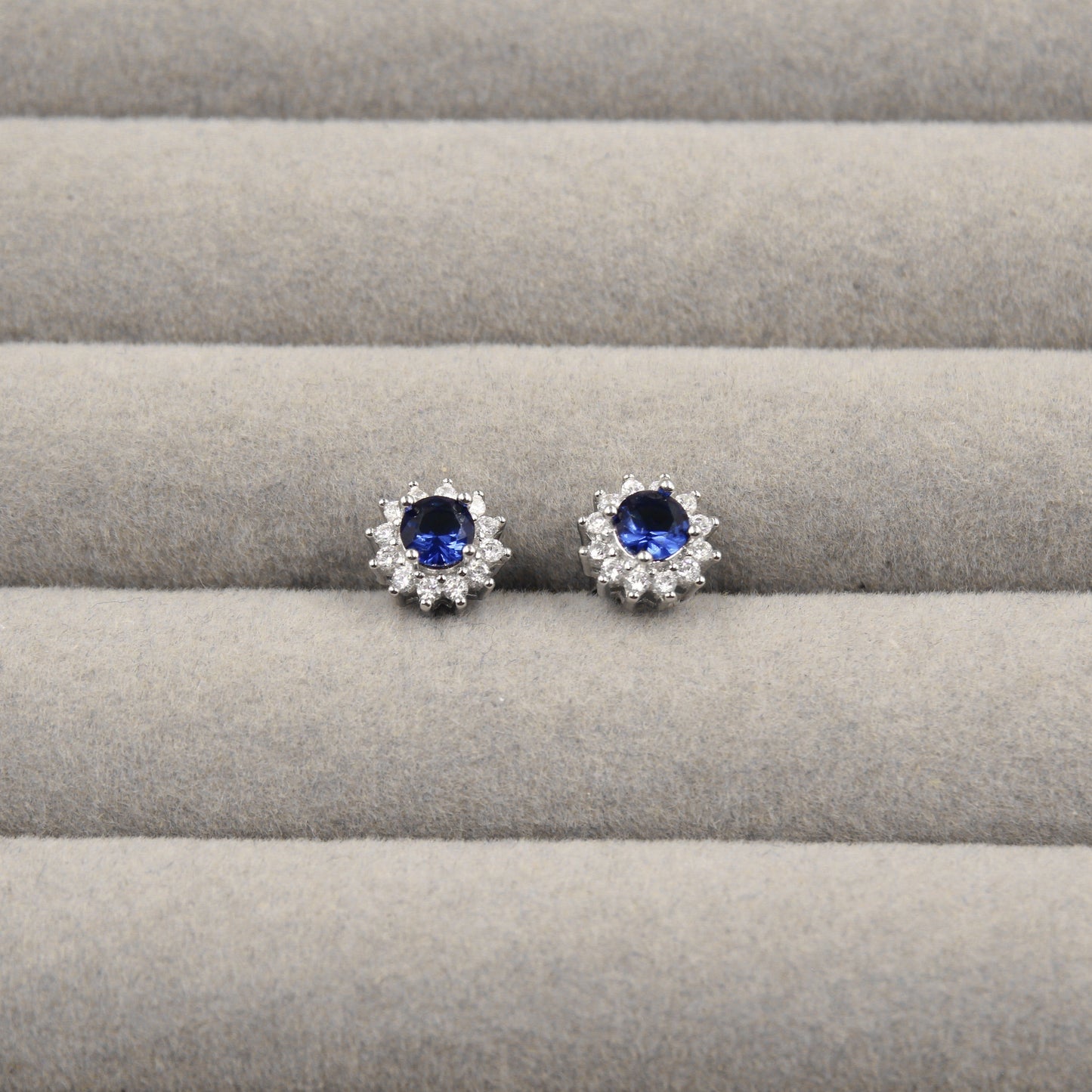 Par de aretes de zafiro azul, rodeados de pequeños diamantes en forma de flor, presentados en un estuche de joyería gris.