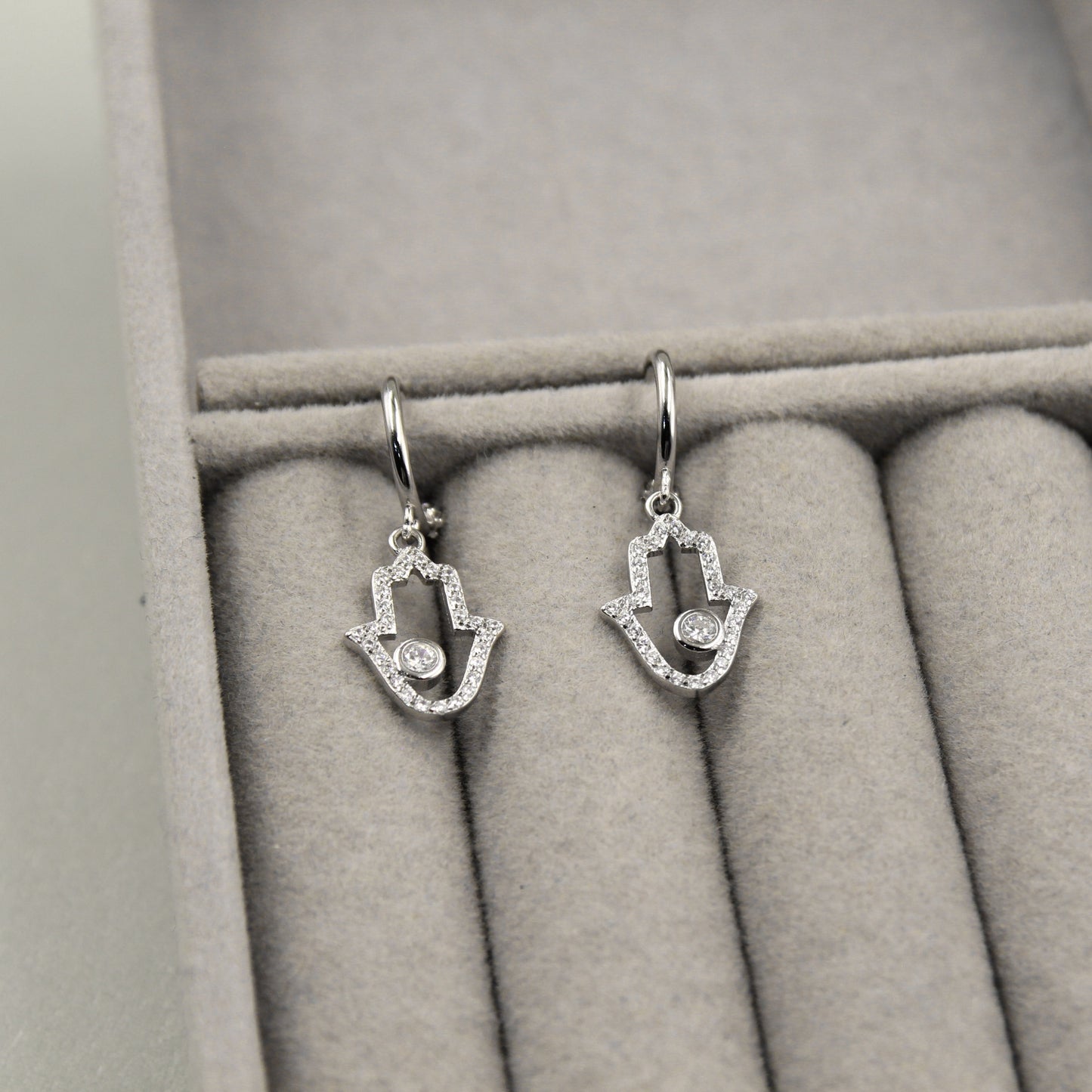 Par de aretes colgantes en forma de mano de Fátima, adornados con pequeños diamantes y un diamante central, presentados en un estuche de joyería gris.