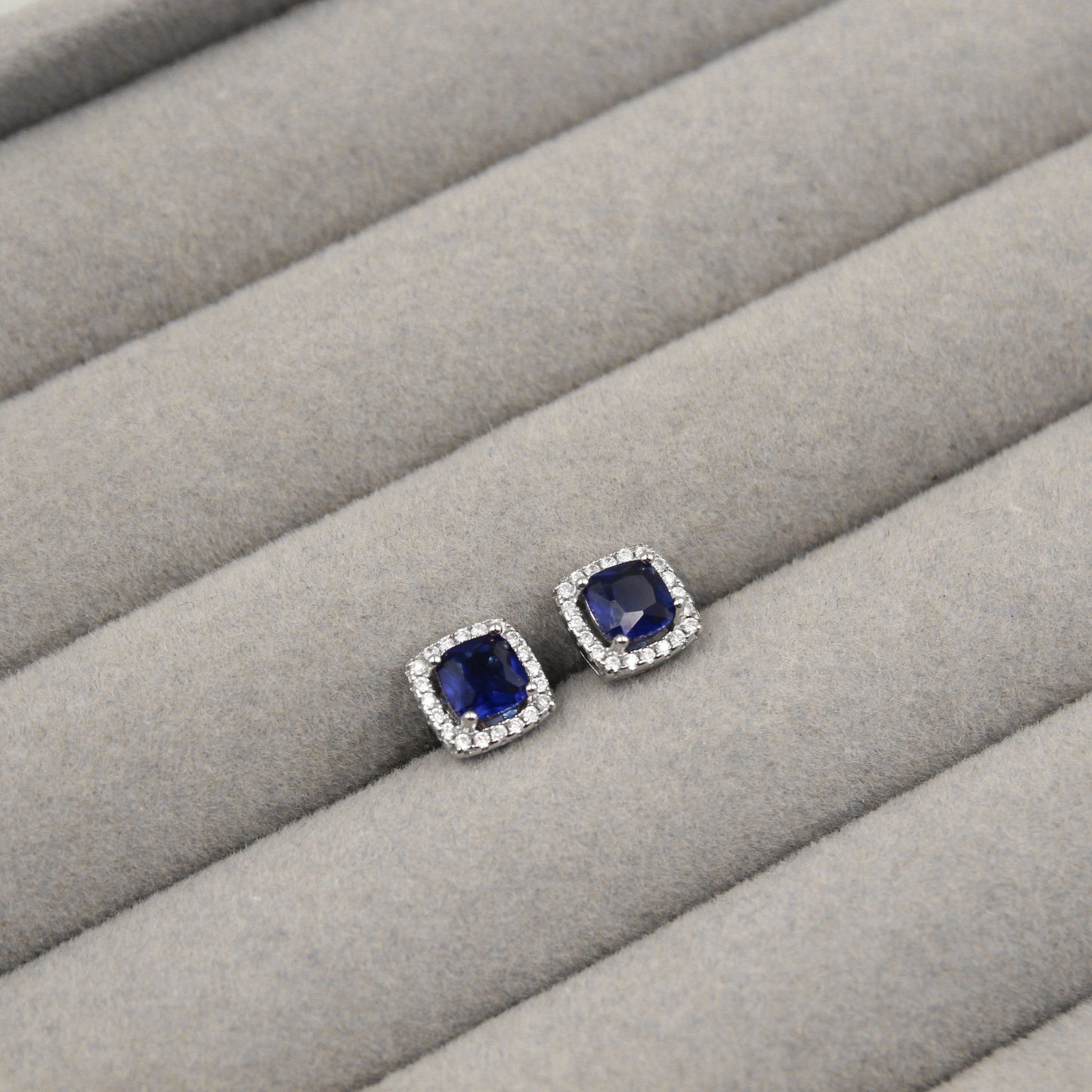 Par de aretes con zafiros azules de corte cuadrado, rodeados de pequeños diamantes, presentados en un estuche de joyería gris.