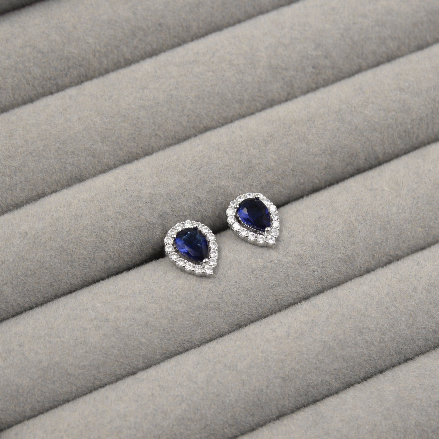 Par de aretes con zafiros azules en forma de lágrima, rodeados de pequeños diamantes, presentados en un estuche de joyería gris.
