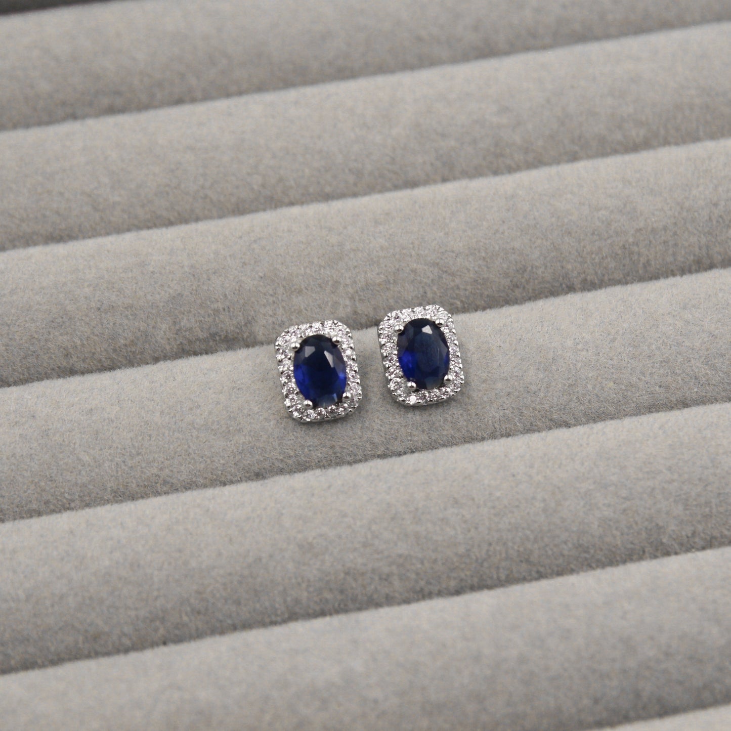 Par de aretes con zafiros azules de corte ovalado, rodeados de pequeños diamantes en un marco rectangular, presentados en un estuche de joyería gris."