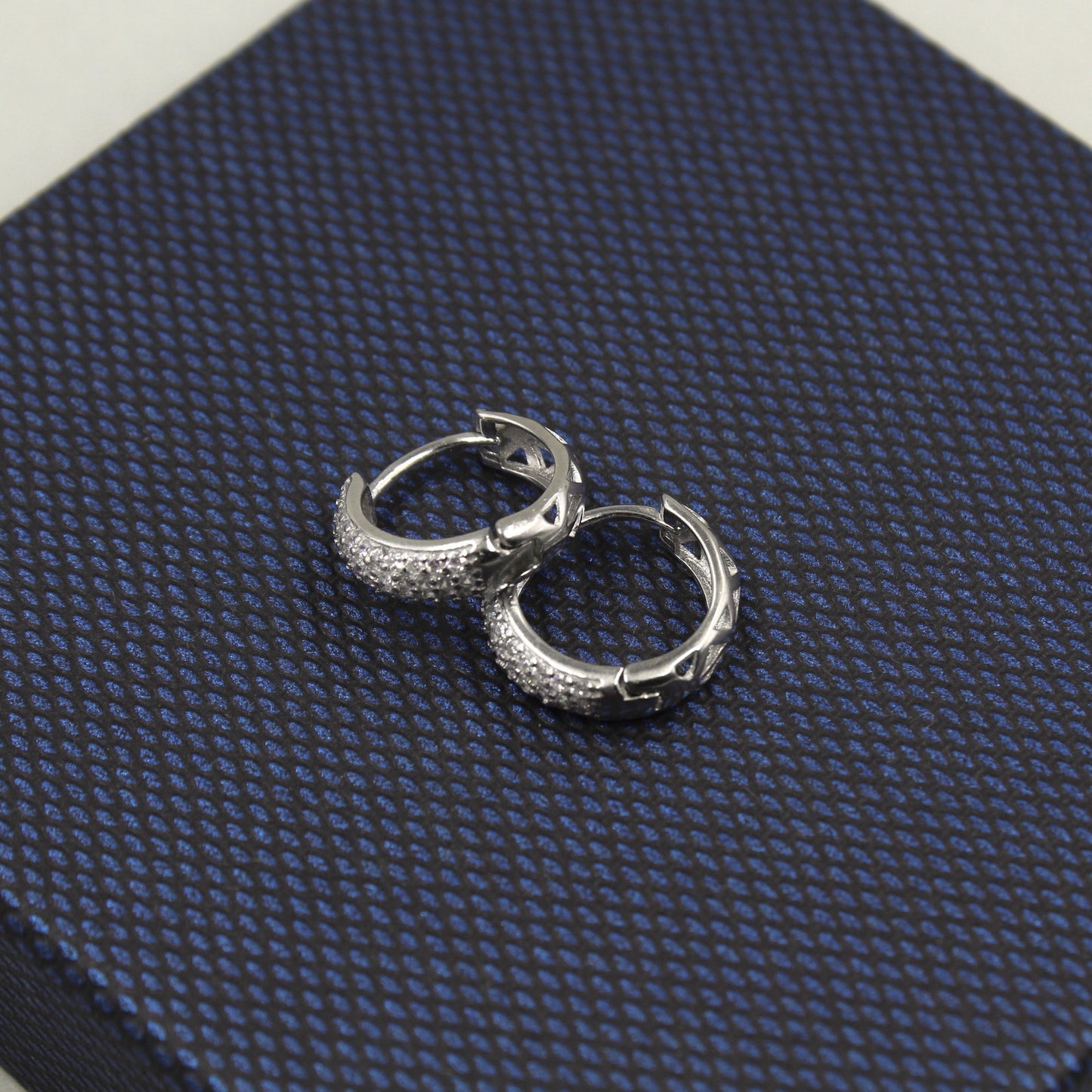 Par de aretes pequeños de aro, completamente cubiertos de diamantes, presentados sobre un fondo texturizado azul oscuro.