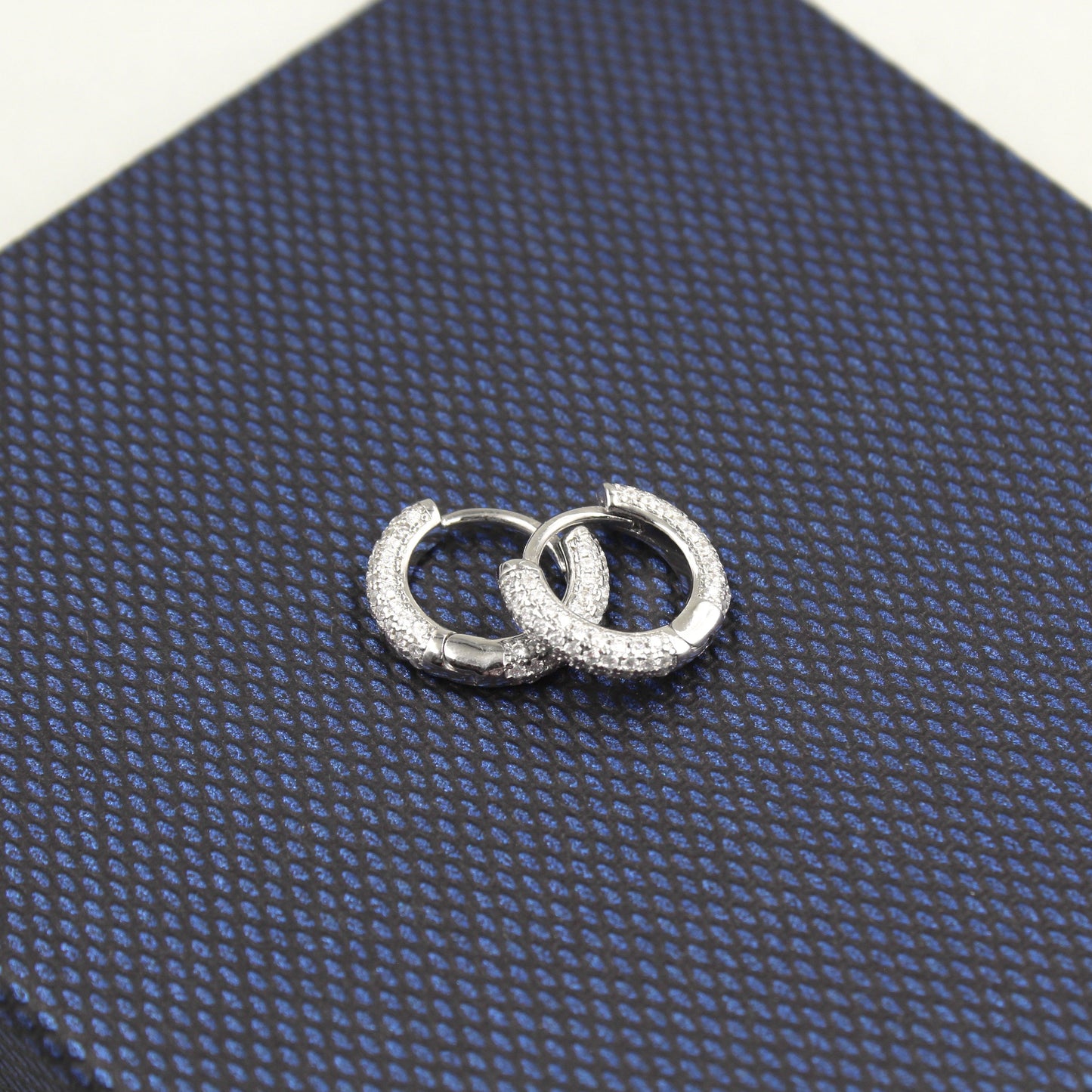 Par de aretes pequeños de aro, completamente cubiertos de diamantes, presentados sobre un fondo texturizado azul oscuro.