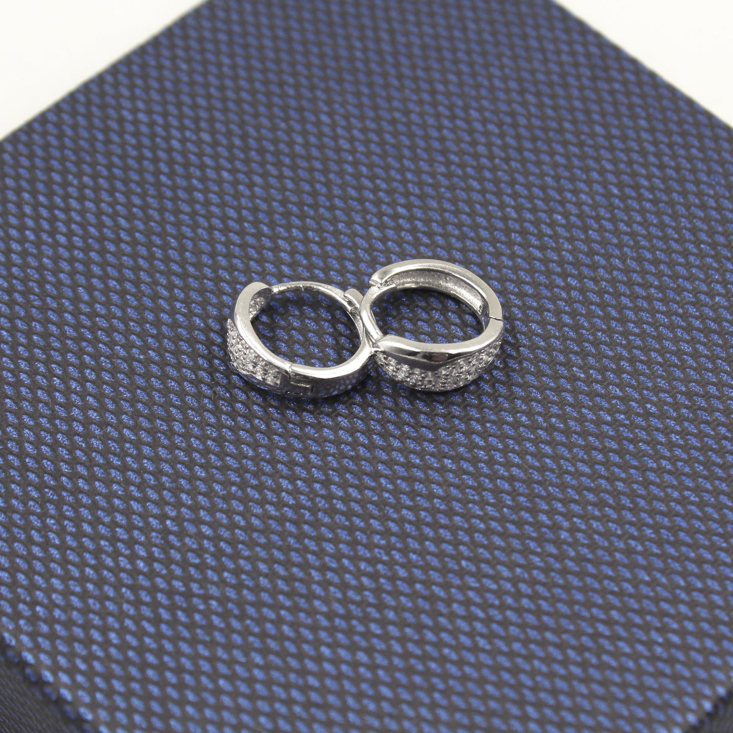 Par de aretes pequeños de aro, adornados con diamantes, presentados sobre un fondo texturizado azul oscuro.