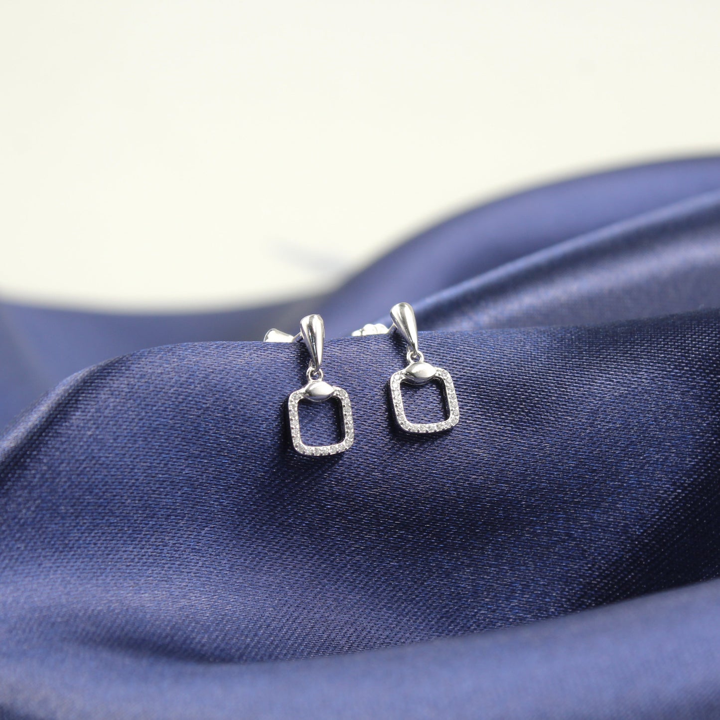 Aretes de plata con forma de rectángulo hueco y borde de diamantes, colocados sobre tela azul.