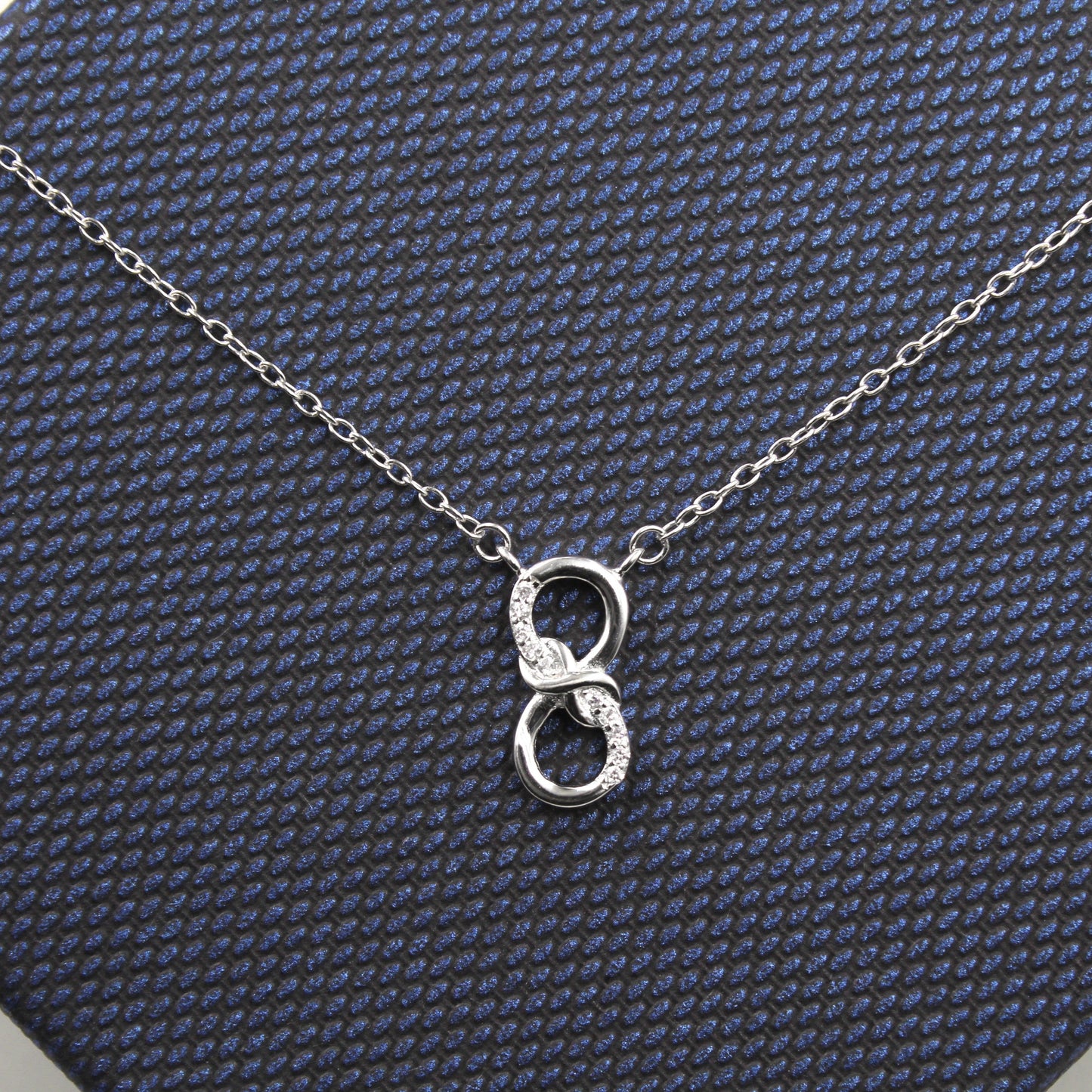 collar de plata con un colgante en forma de dos aros entrelazados sobre una caja de regalo con textura azul oscuro. El aro superior está parcialmente decorado con pequeñas circonias cúbicas que agregan un toque brillante, mientras que el aro inferior es de plata lisa.