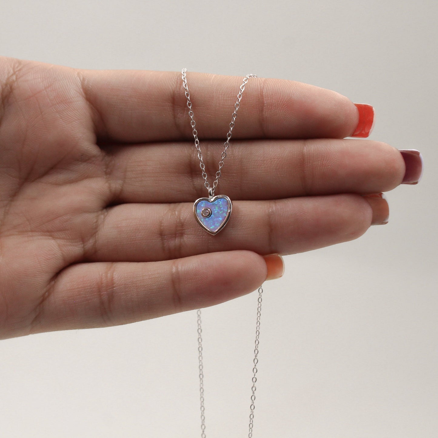 mano sosteniendo un delicado collar de plata con un colgante en forma de corazón de madreperla azul. El corazón refleja variaciones sutiles de azul y blanco, proporcionando un brillo y una profundidad únicos. La cadena de eslabones finos complementa elegantemente el colgante.