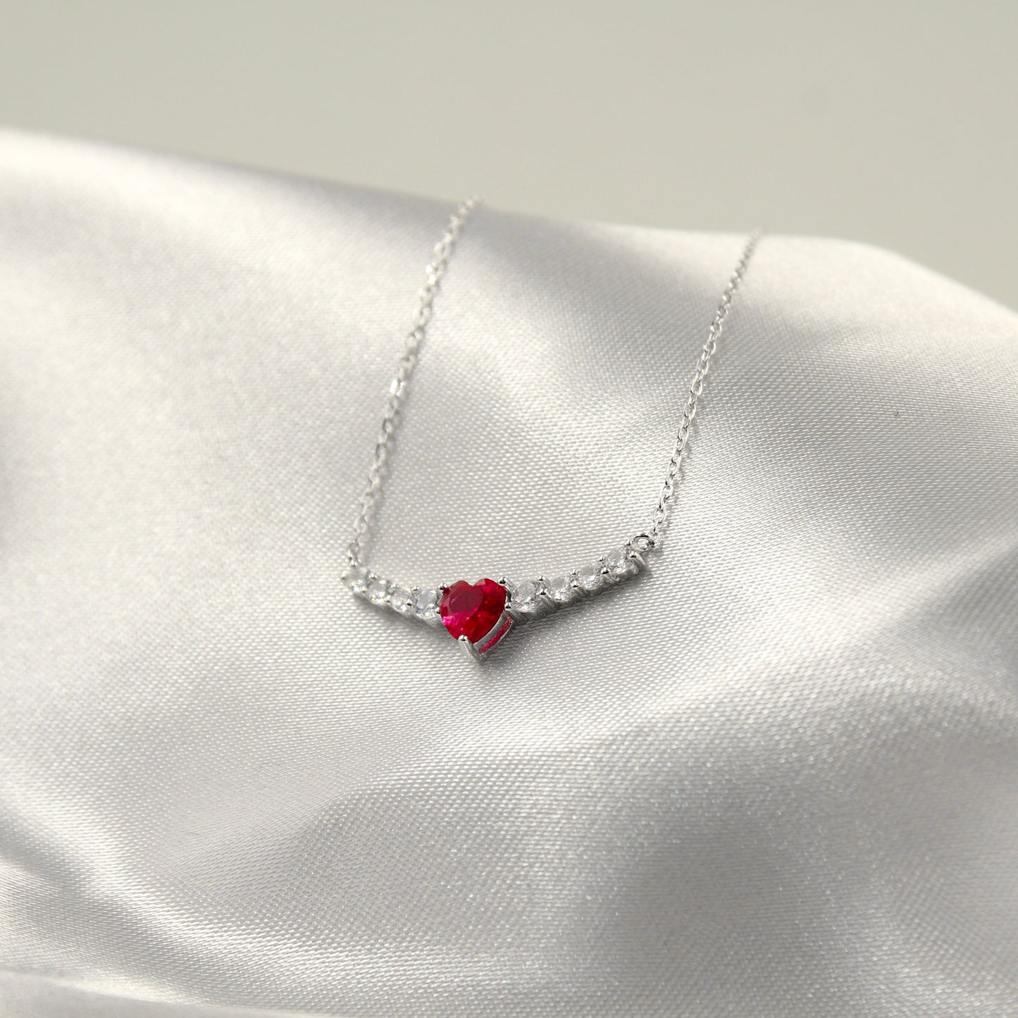 delicado collar de plata con un colgante en forma de corazón que presenta una vibrante piedra roja en el centro, rodeada por pequeñas circonias cúbicas que añaden un toque de brillo. El collar se dispone sobre una tela de satén blanco, lo que resalta su colorido y elegancia.
