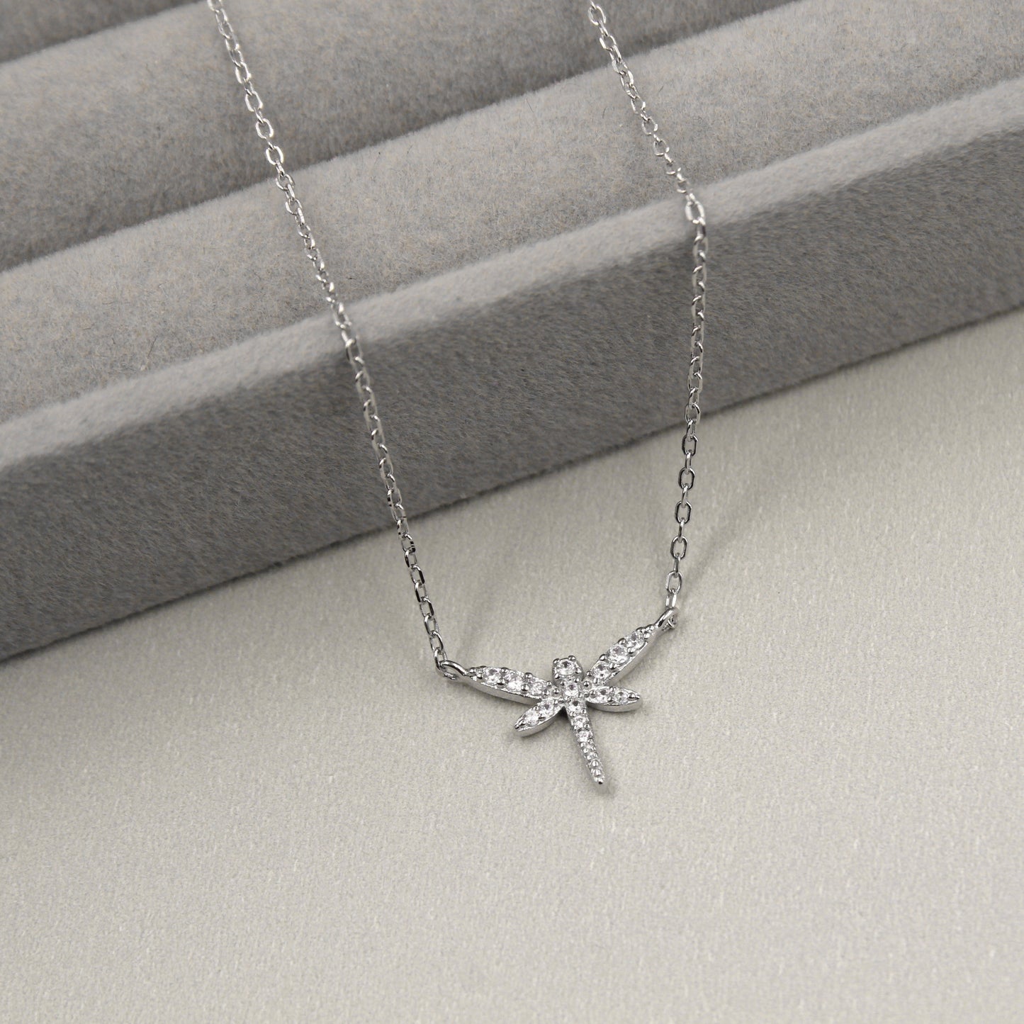 delicado collar de plata con un colgante en forma de libélula, cuyas alas y cuerpo están elegantemente adornados con pequeñas circonias cúbicas que proporcionan un brillo sutil. El colgante se presenta sobre un fondo de tela gris con líneas horizontales suaves.