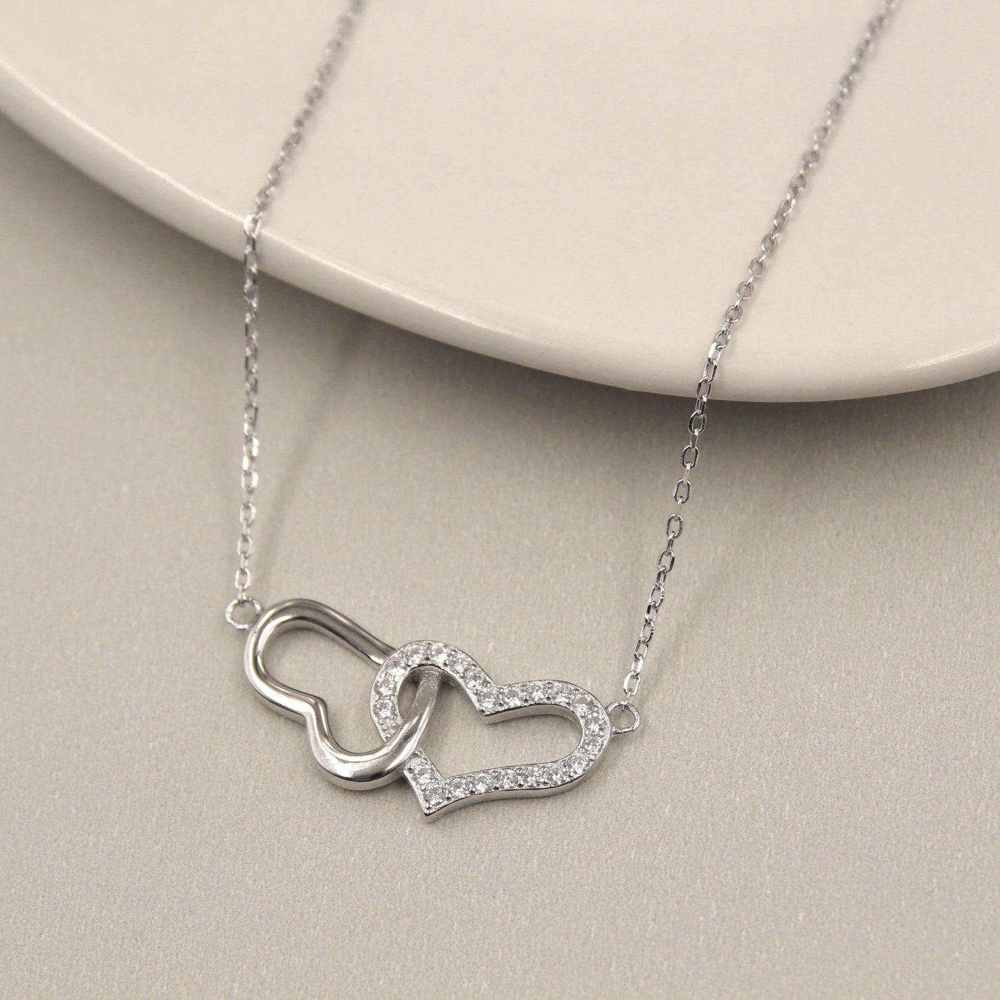 collar de plata con dos colgantes de corazón entrelazados. Uno de los corazones es liso y de plata sólida, mientras que el otro está cubierto con circonias cúbicas brillantes, ofreciendo un contraste elegante. El collar descansa sobre una placa de cerámica de color claro que realza su diseño y brillo.