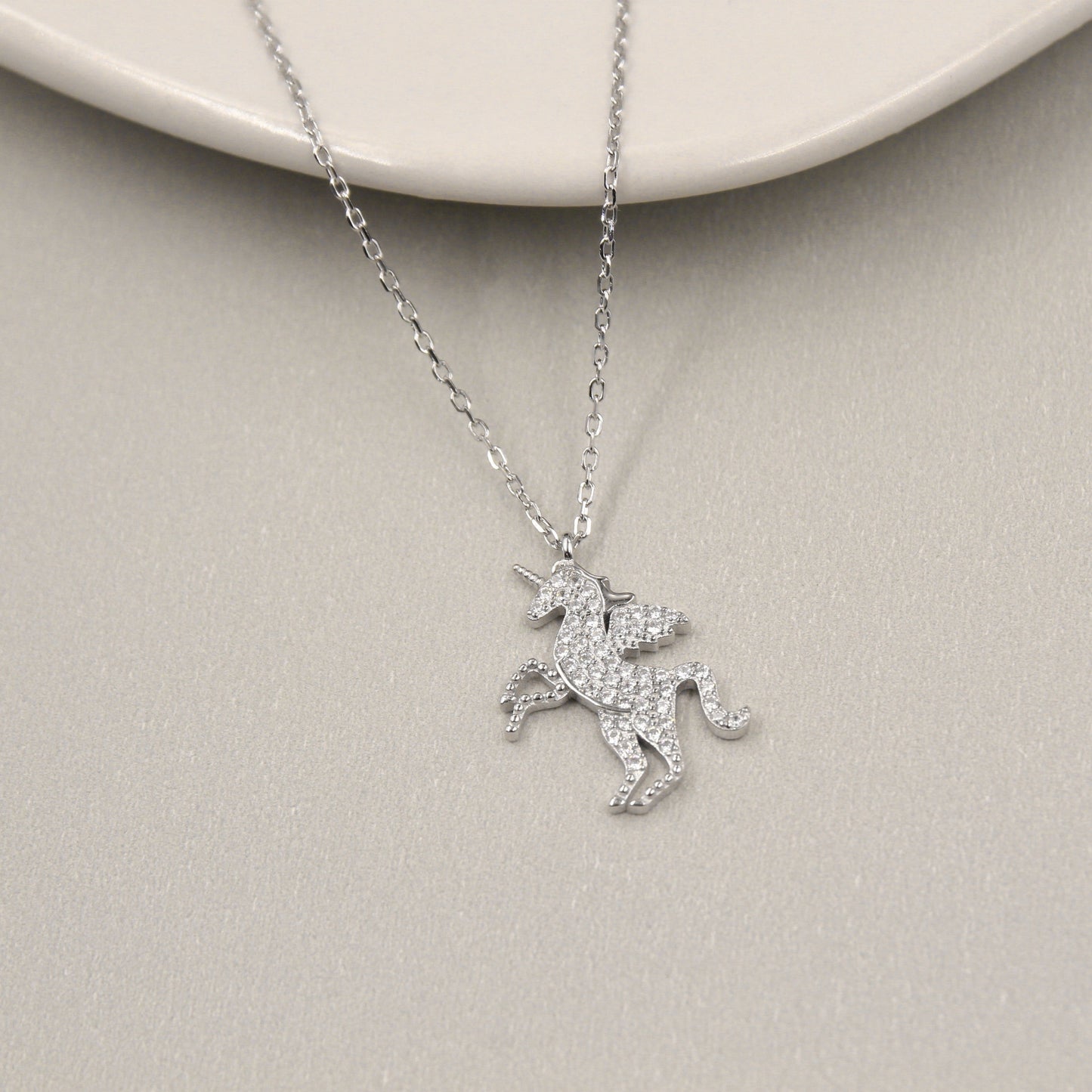 collar de plata con un colgante en forma de unicornio. El unicornio está adornado con circonias cúbicas que cubren todo su cuerpo y cuerno, ofreciendo un brillo excepcional. El colgante se presenta sobre un fondo de cerámica blanca que realza su detalle y luminosidad.