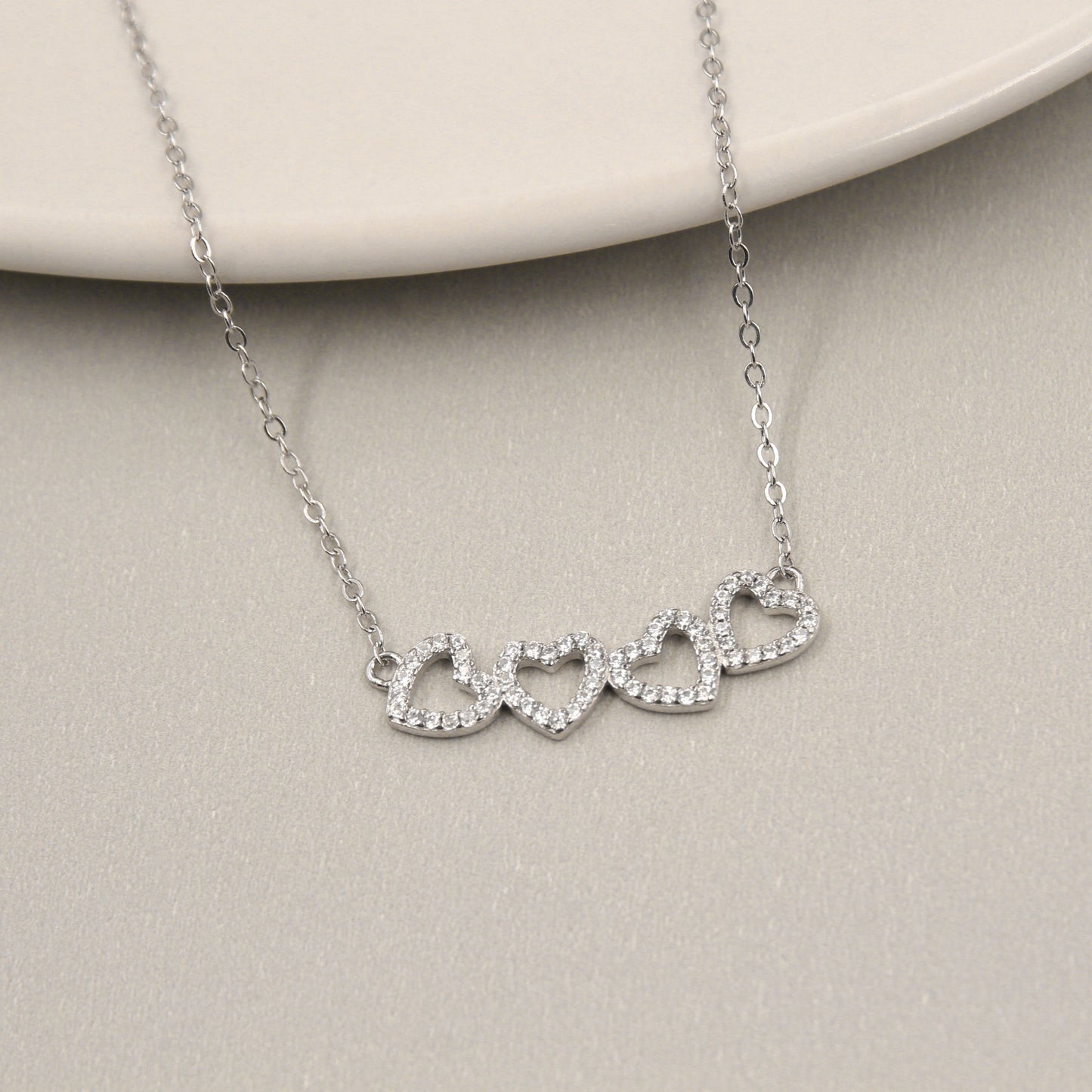 delicado collar de plata con un diseño de cuatro corazones entrelazados. Cada corazón está contorneado por pequeñas circonias cúbicas que brillan intensamente. El collar está dispuesto sobre un plato de cerámica de color claro, añadiendo un contraste suave y elegante al brillante metal.