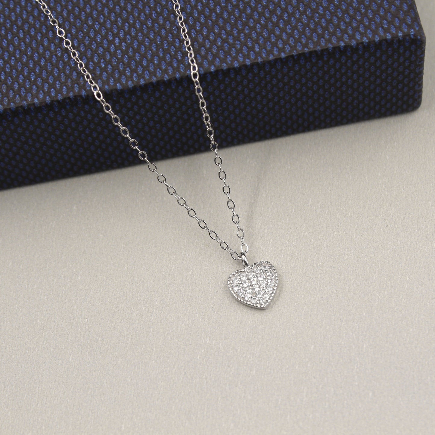 elegante collar de plata con un colgante en forma de corazón completamente cubierto de circonias cúbicas brillantes. El colgante está suspendido de una cadena de eslabones finos, presentado sobre una caja de regalo de color azul oscuro con textura, destacando su brillo y diseño romántico.