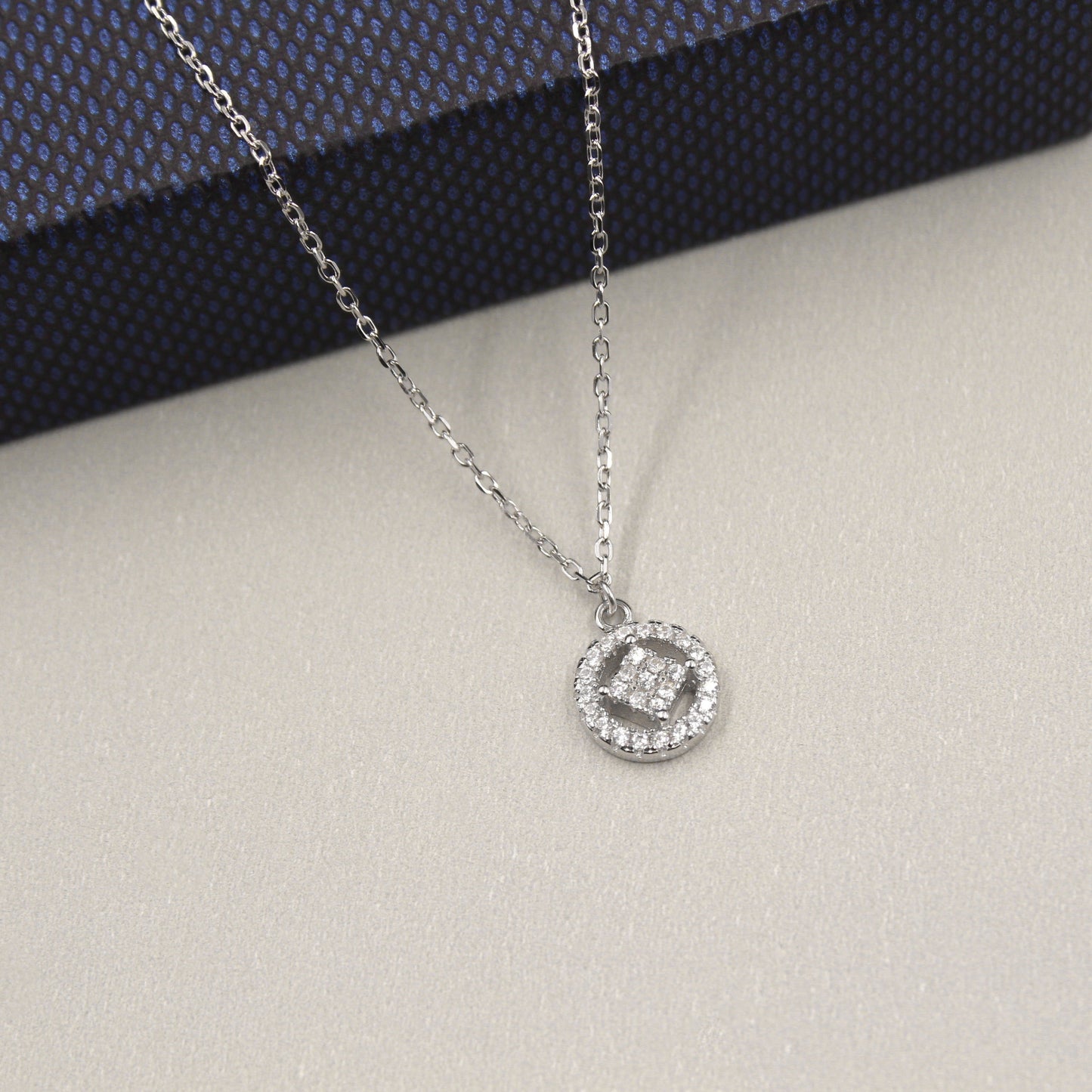 collar de plata elegante con un colgante circular. El colgante tiene un diseño entrelazado en el centro, embellecido con pequeñas circonias cúbicas que le dan un brillo sutil. La cadena es delgada y de eslabones finos. El collar está sobre un fondo gris claro con una caja de regalo de color azul oscuro con textura en el fondo.