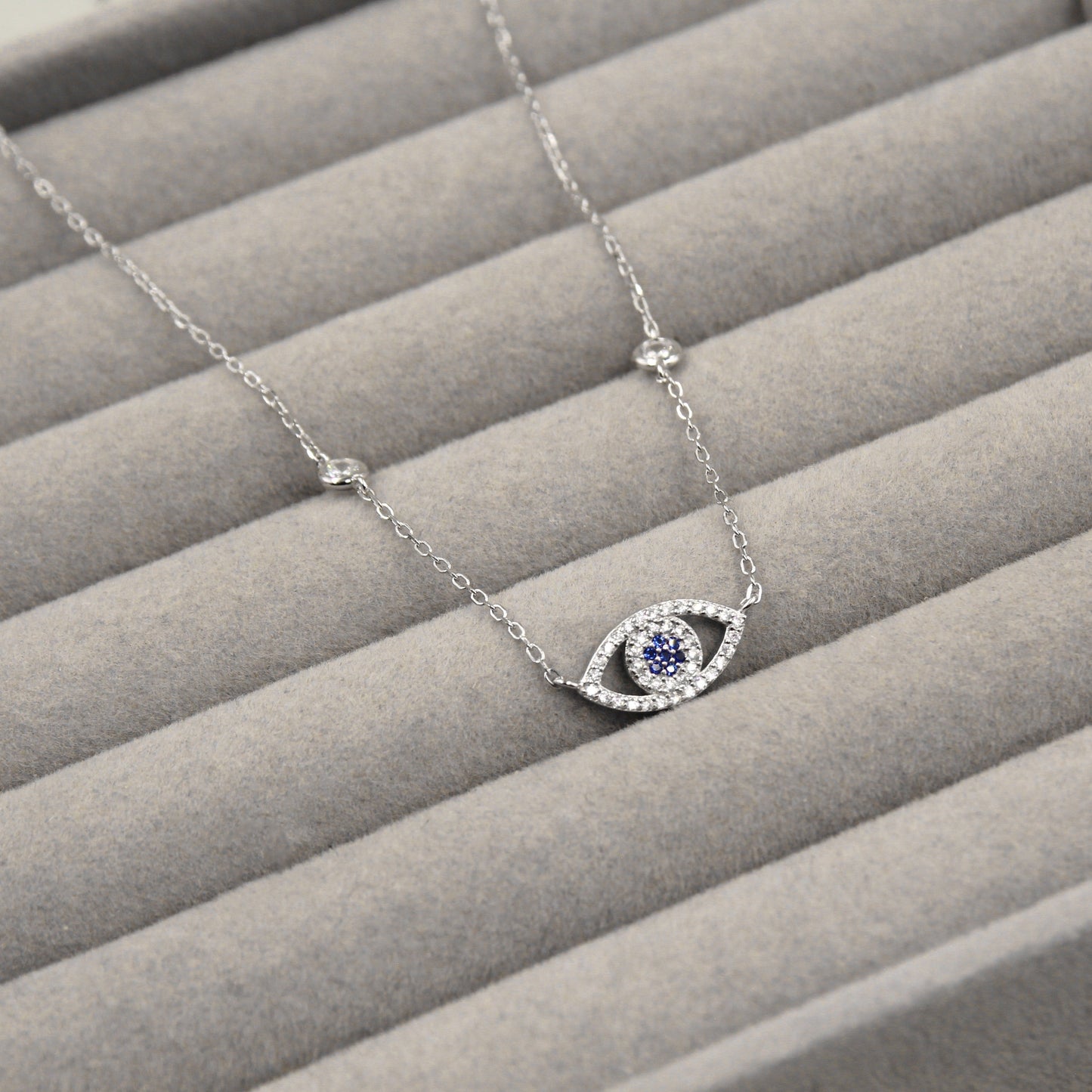 collar de plata con un colgante en forma de ojo, destacando una piedra azul en el centro rodeada por un halo de circonias cúbicas. El diseño evoca protección y estilo, presentado sobre un fondo de terciopelo gris que realza su brillo y color vibrante.