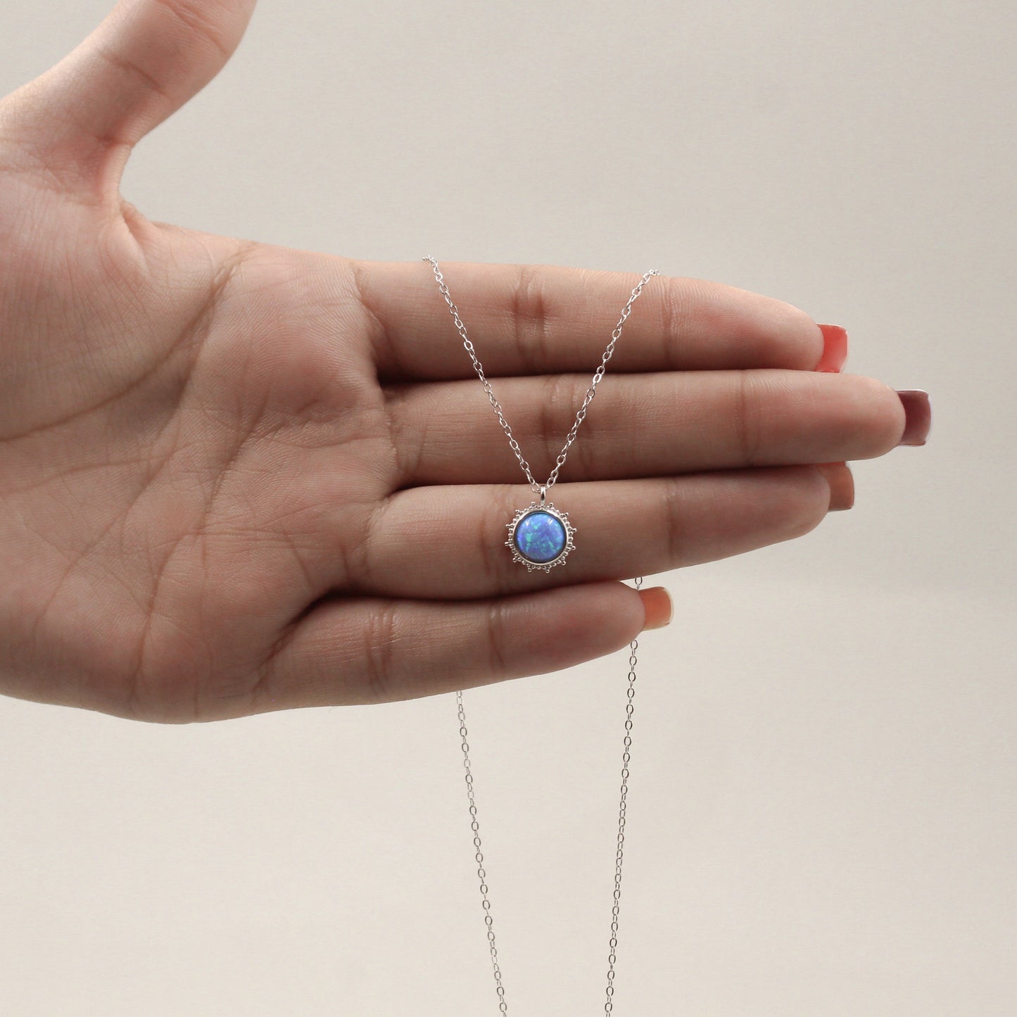 una mano sosteniendo un collar de plata con un colgante redondo que alberga una piedra de ópalo azul vibrante, rodeada por un halo de pequeñas circonias cúbicas. El diseño capta la luz, mostrando destellos de colores dentro del ópalo, mientras la cadena delicada se extiende elegantemente desde ambos lados del colgante.