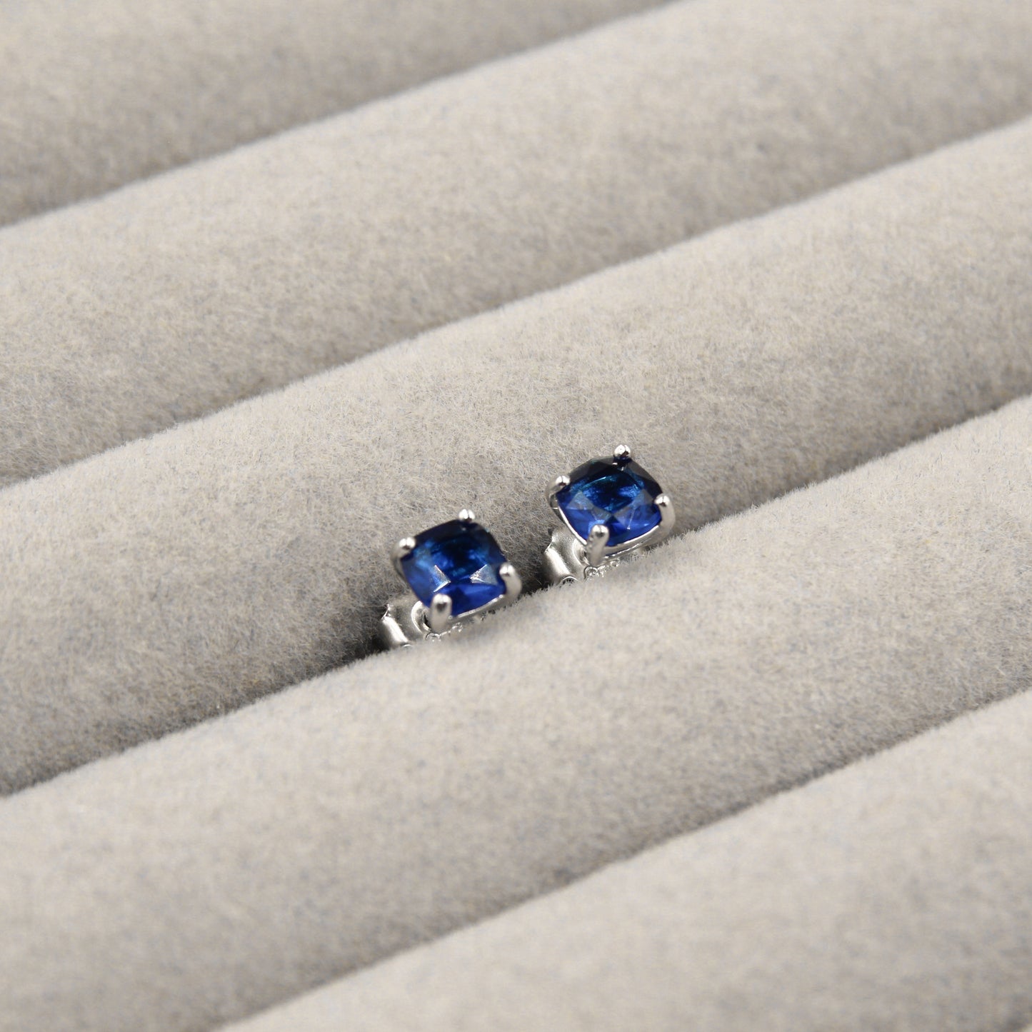 Par de aretes pequeños con zafiros azules cuadrados, presentados en un estuche de joyería gris.