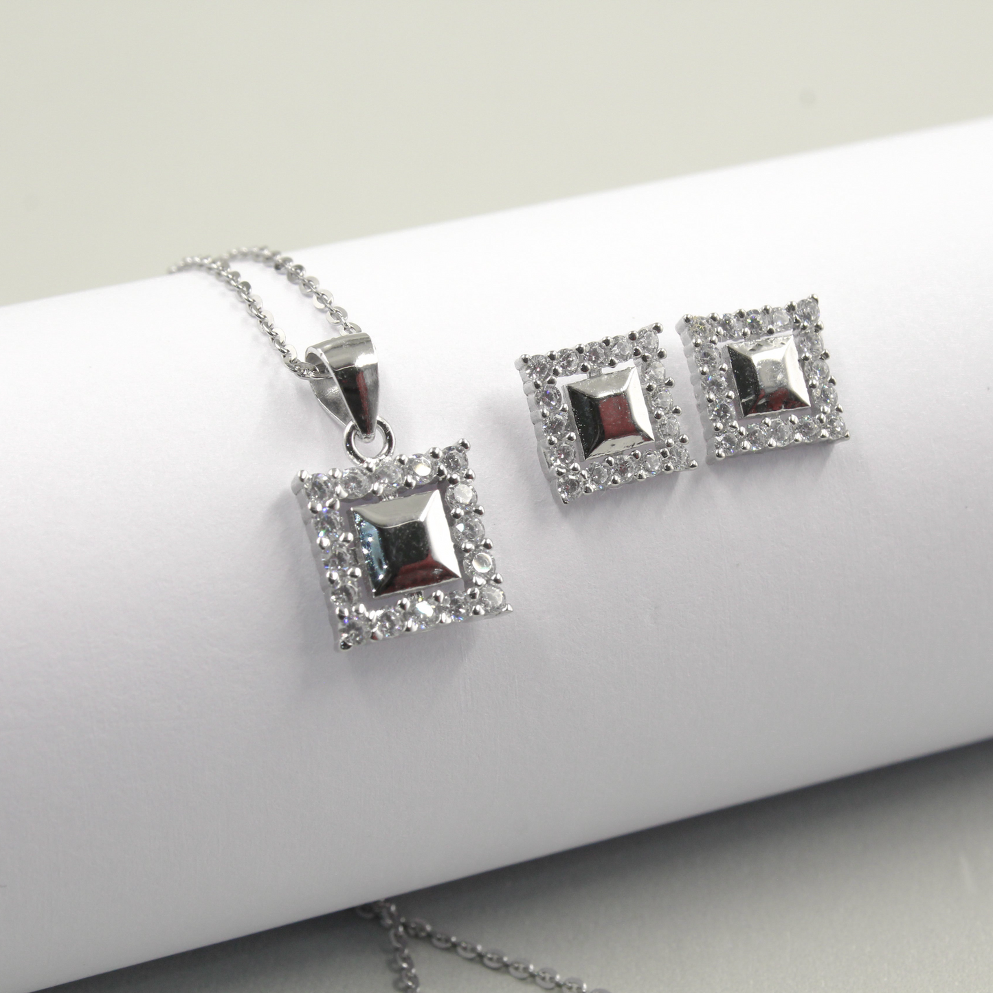 Juego de joyería con colgante y aretes cuadrados decorados con pequeños diamantes, sobre fondo blanco."