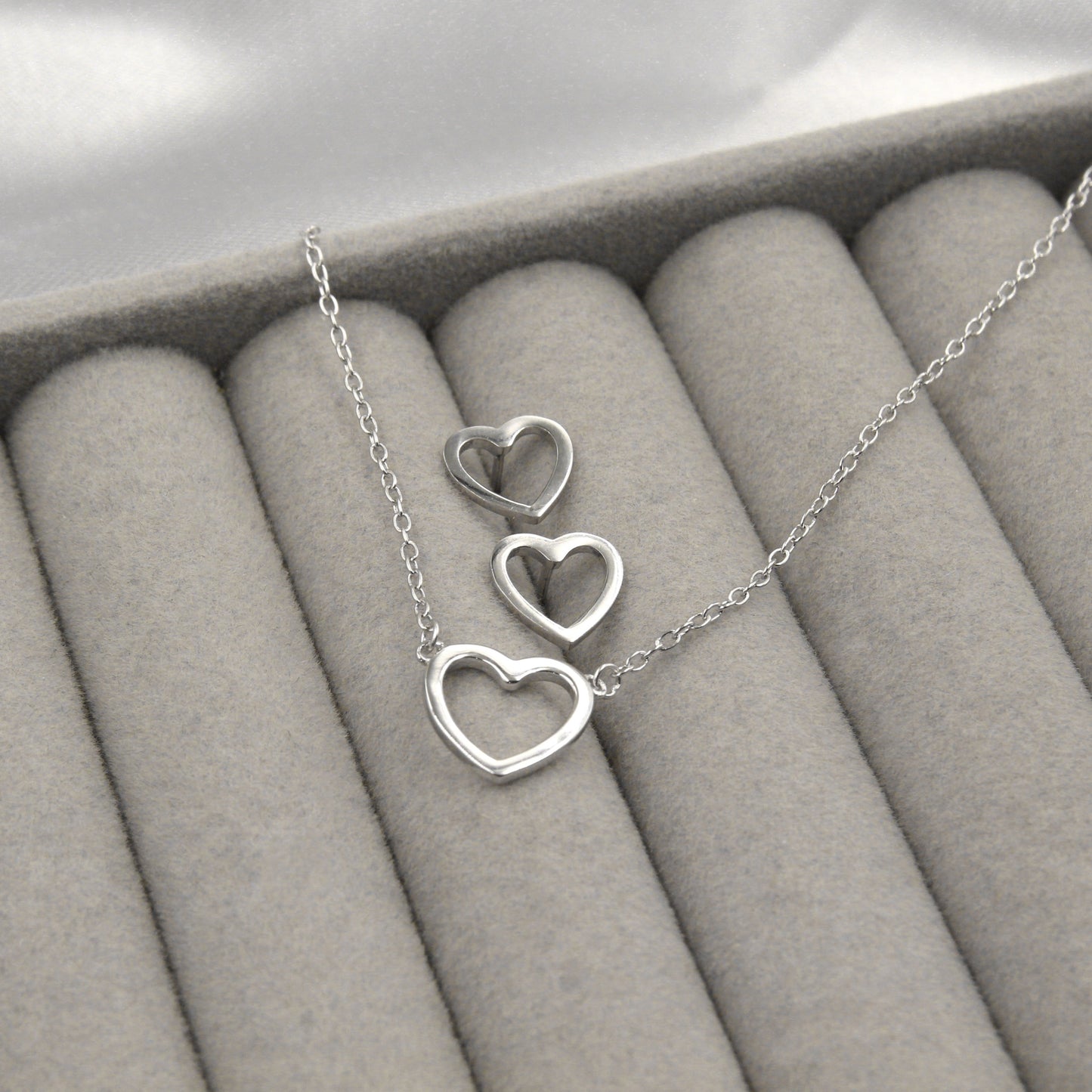 Juego de joyería con colgante y aretes en forma de corazón, hechos de plata, presentados en una caja de joyería gris, sobre fondo blanco.