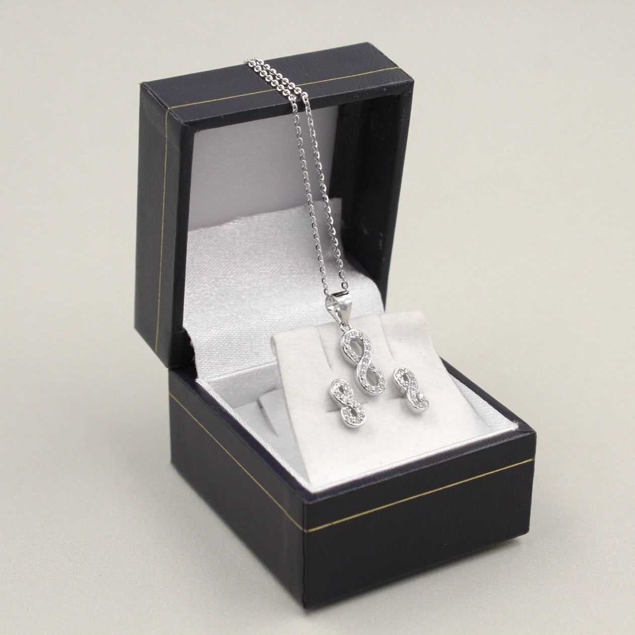 Juego de joyería con colgante y aretes en forma de infinito, decorados con pequeños diamantes, presentado en una caja negra.