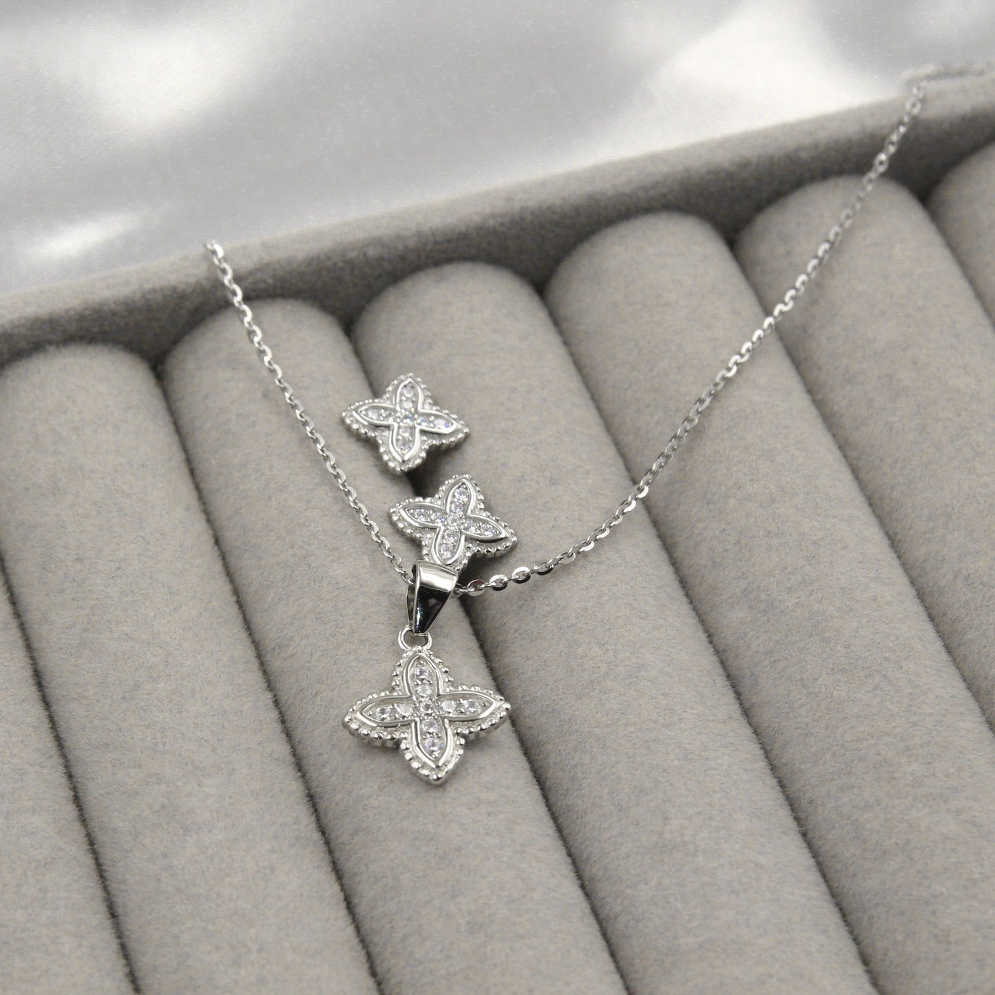 Juego de joyería con colgante y aretes en forma de flor, decorados con pequeños diamantes, presentados en una caja de joyería gris.