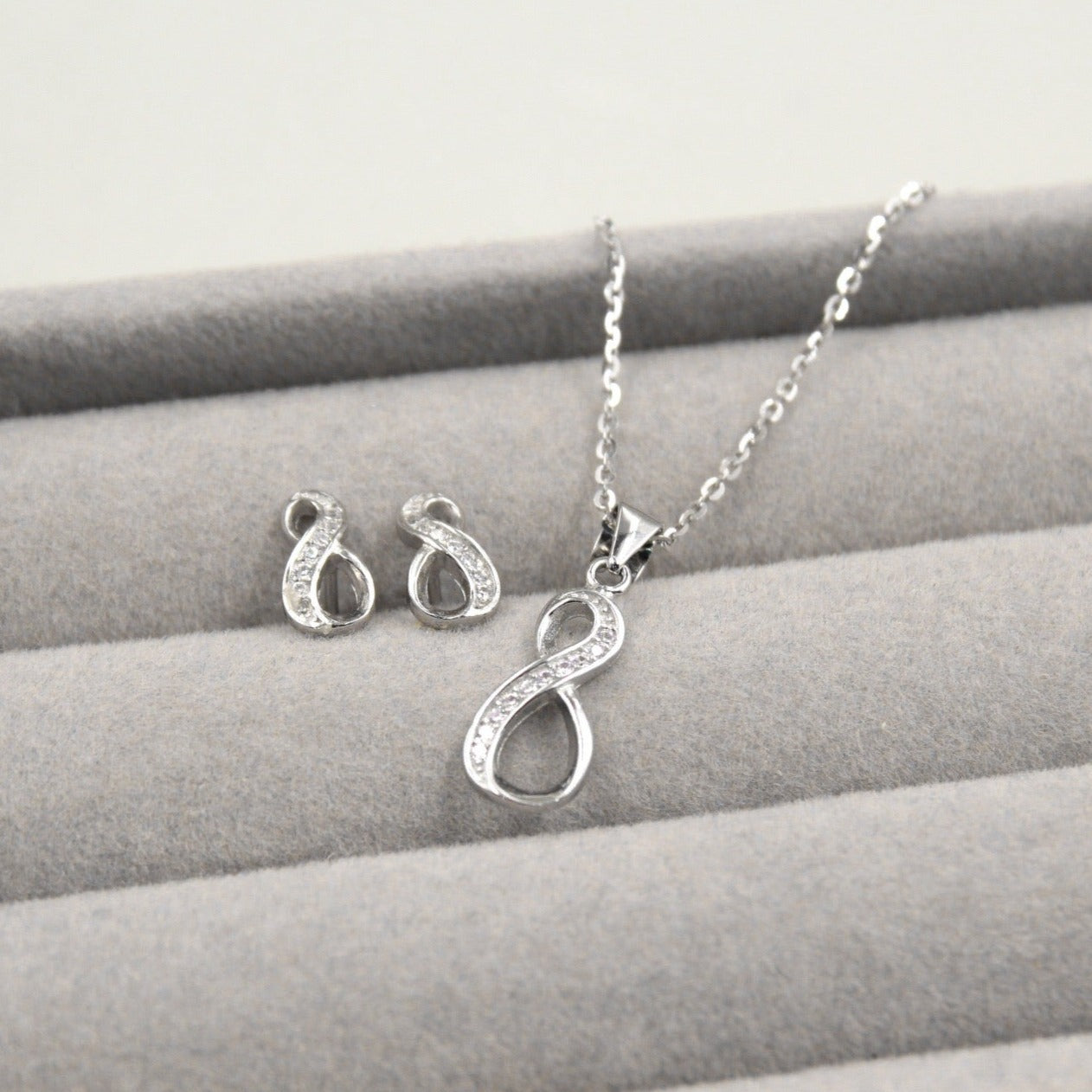 Juego de joyería con colgante y aretes en forma de infinito, decorados con pequeños diamantes, presentados en una caja de joyería gris.