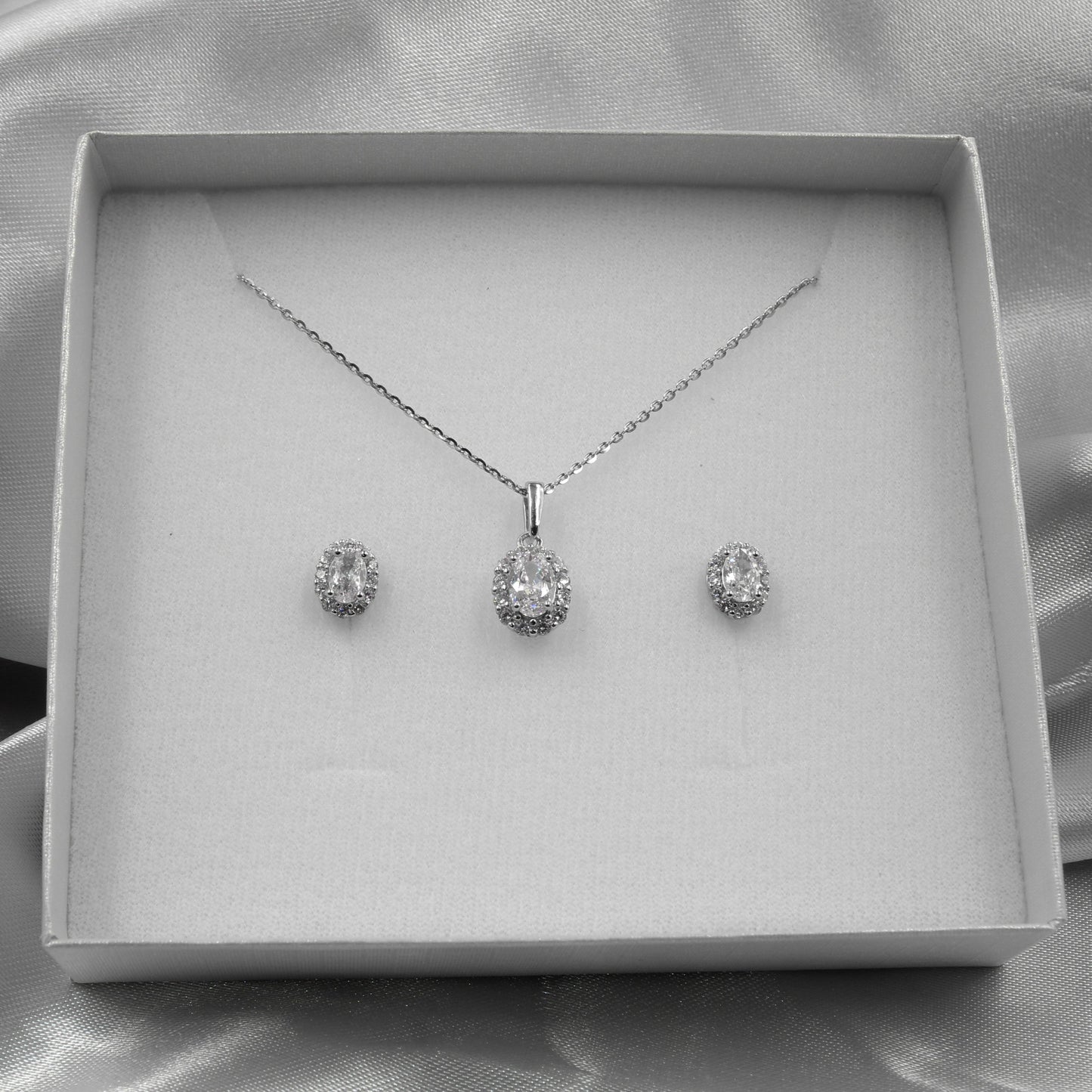 Juego de joyería con colgante y aretes ovalados, decorados con pequeños diamantes, presentados en una caja blanca sobre fondo de tela plateada.