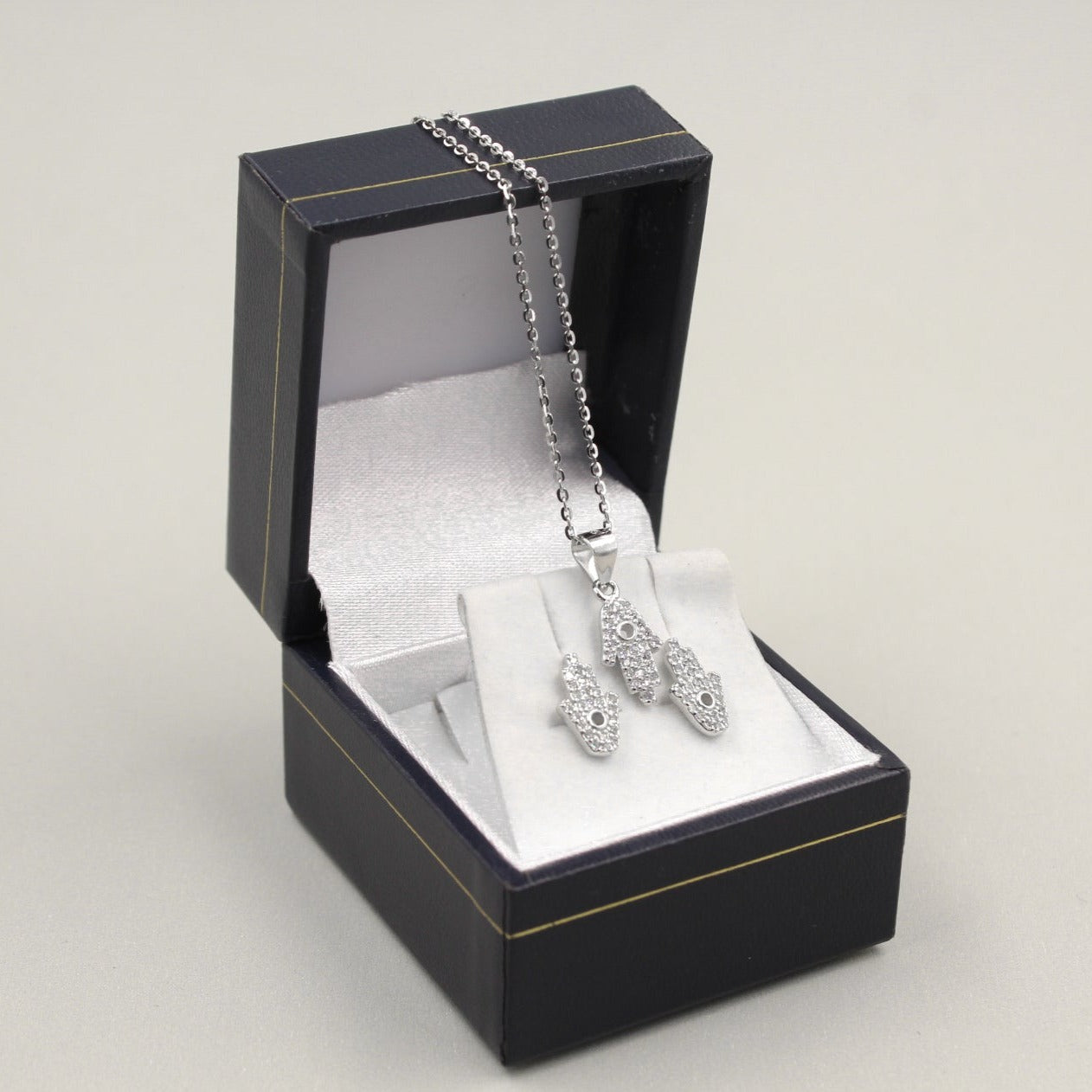 Juego de joyería con colgante y aretes en forma de mano de Fátima, decorados con pequeños diamantes, presentados en una caja negra.