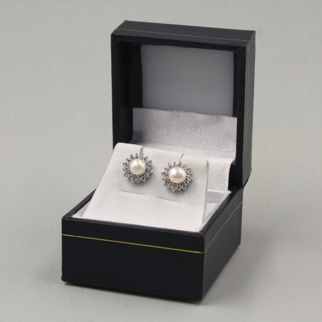 par de aretes de plata en una caja de joyería negra con detalles dorados. Cada arete presenta una perla central blanca rodeada por un halo de pequeñas circonias cúbicas, lo que les da un aspecto elegante y clásico.
