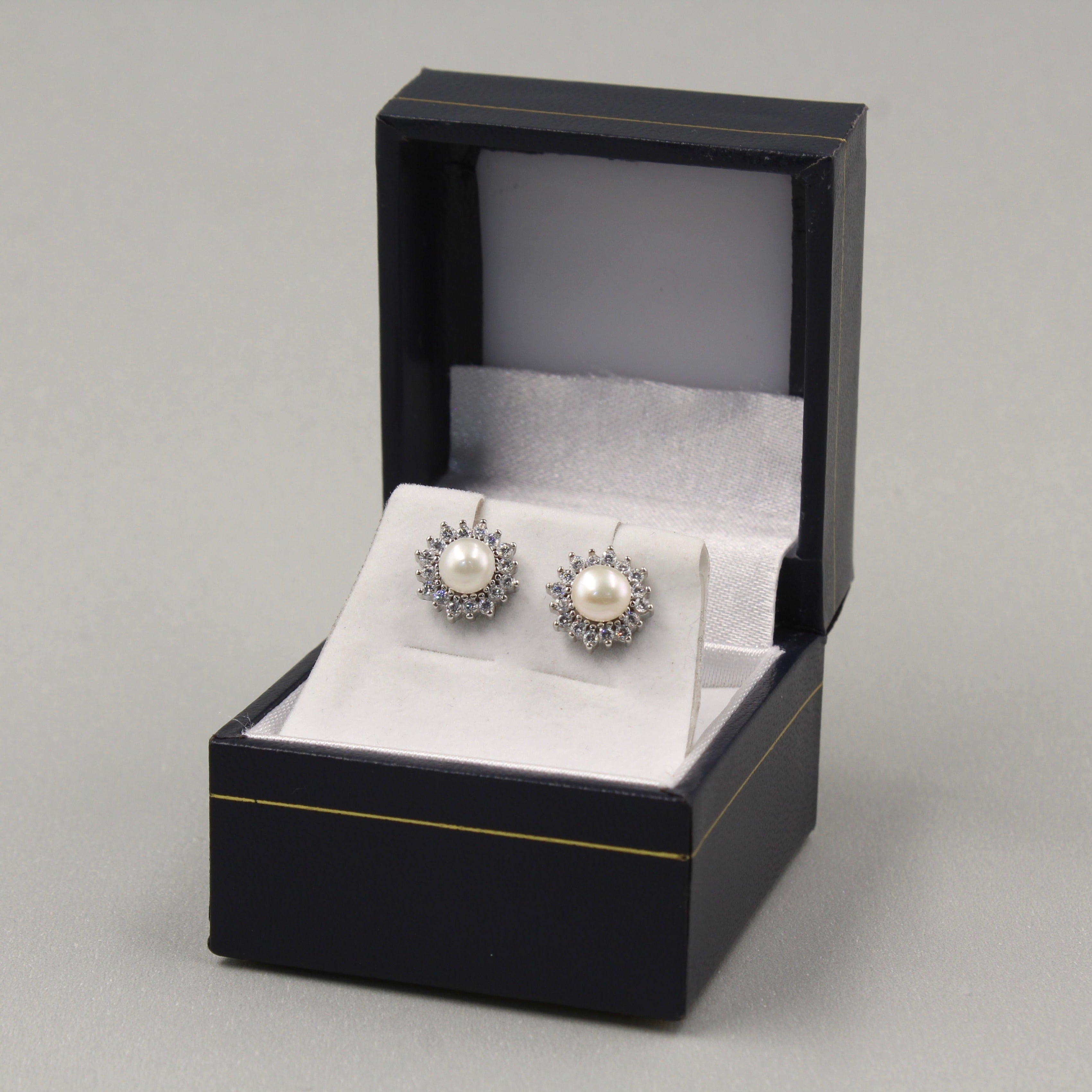 par de aretes de plata en una caja de joyería negra con detalles dorados. Cada arete presenta una perla central blanca rodeada por un halo de pequeñas circonias cúbicas, lo que les da un aspecto elegante y clásico.