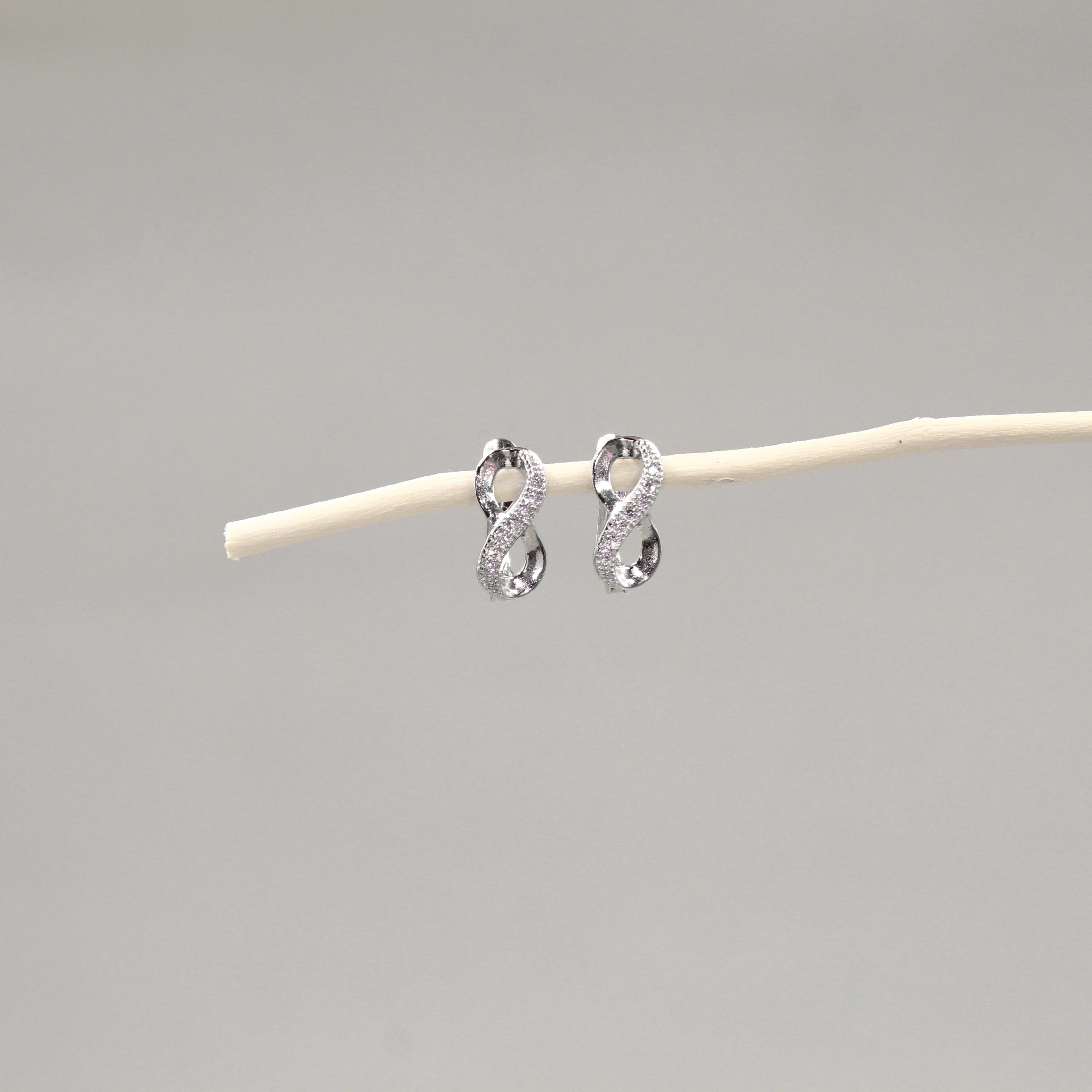 Dos pendientes de plata en forma de lazo infinito con incrustaciones de cristales, colocados delicadamente sobre una varilla de madera pálida, con un fondo gris claro que realza su diseño brillante y elegante.