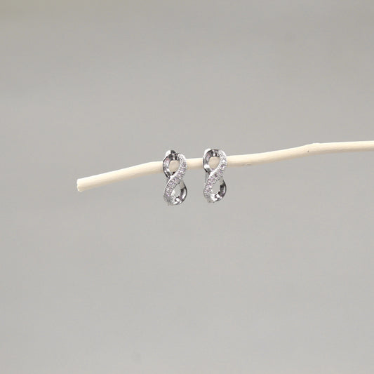 Dos pendientes de plata en forma de lazo infinito con incrustaciones de cristales, colocados delicadamente sobre una varilla de madera pálida, con un fondo gris claro que realza su diseño brillante y elegante.