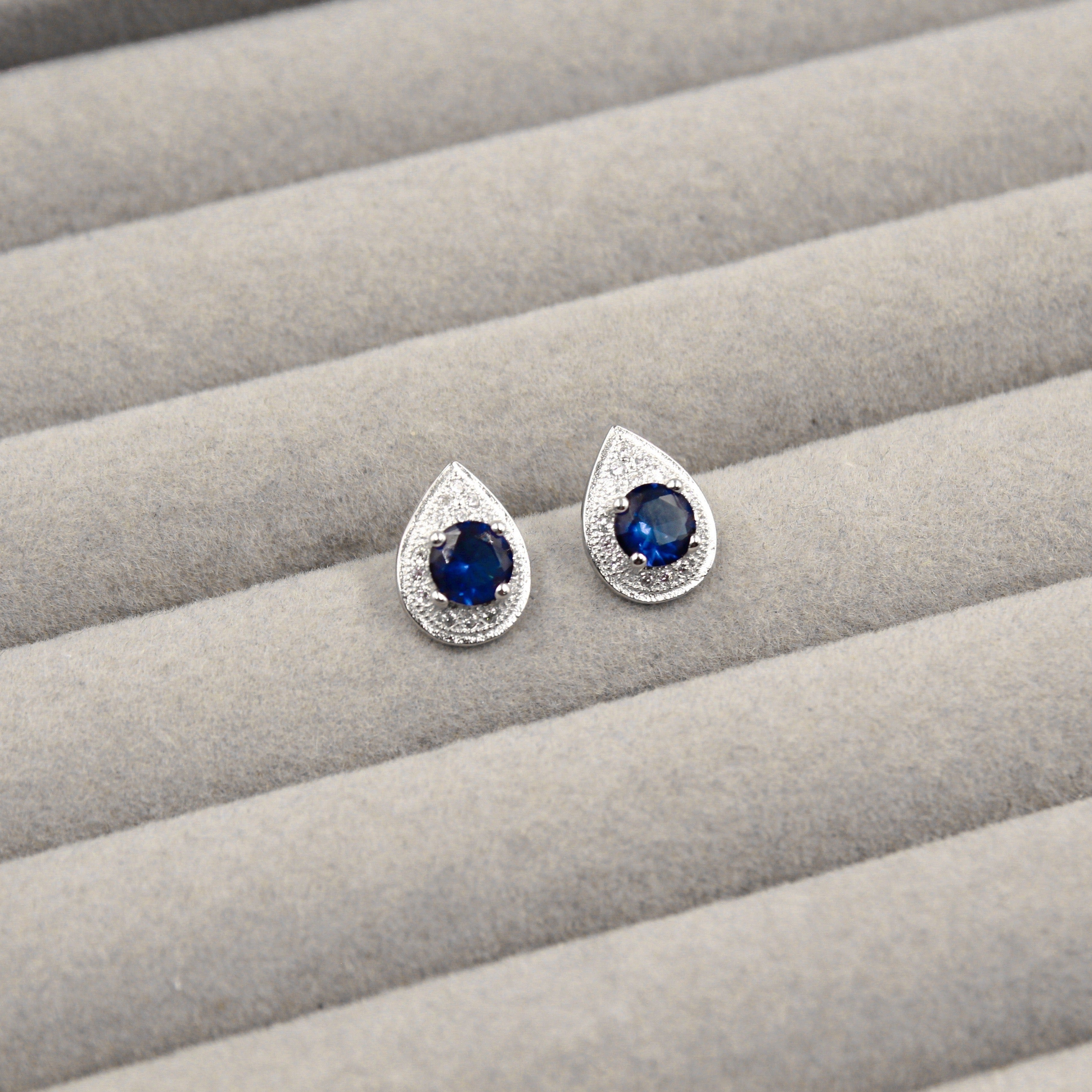 Par de aretes con zafiros azules en forma redonda, montados en un marco en forma de lágrima adornado con pequeños diamantes, presentados en un estuche de joyería gris.