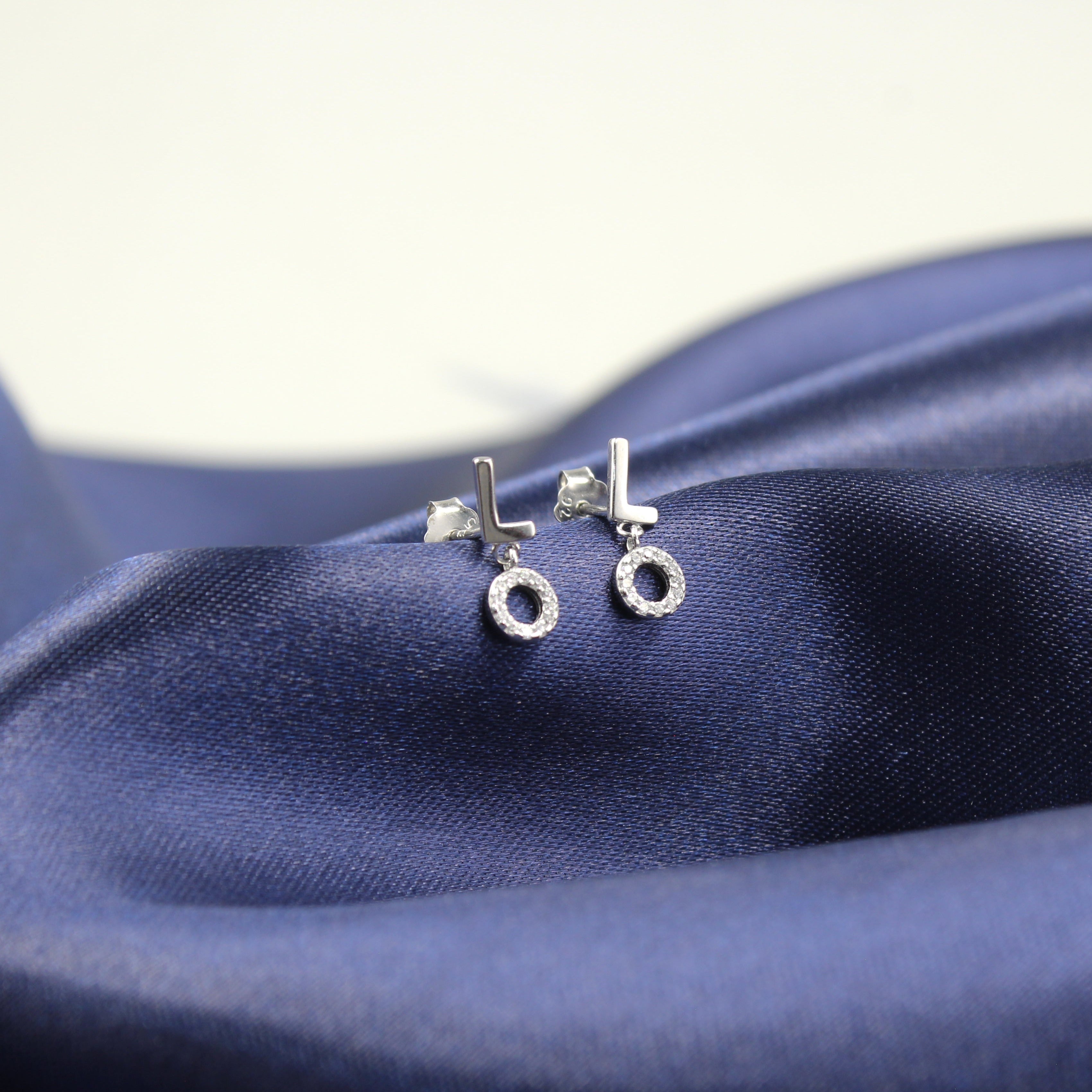 par de aretes de plata con diseño moderno, presentando la palabra 'LOVE' dividida entre ambos. Las letras 'L' y 'O' en un arete y las letras 'V' y 'E' en el otro, cada letra acompañada por un pequeño círculo de circonias cúbicas. Los aretes están colocados sobre un fondo de tela azul satinada, resaltando su brillo y diseño contemporáneo.