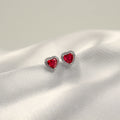 par de aretes en forma de corazón con piedras preciosas rojas rodeadas de diminutos diamantes en una superficie de satén blanco.