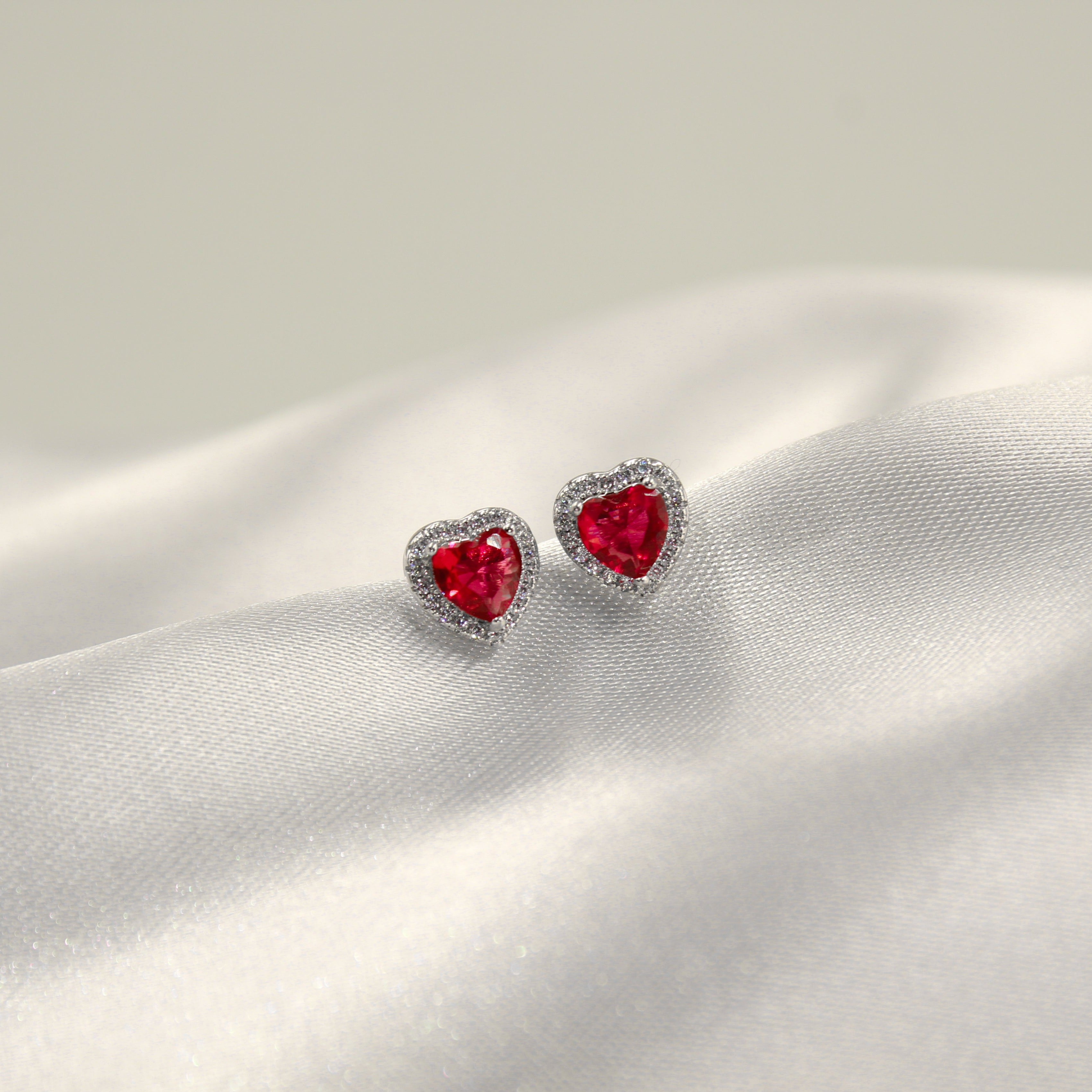 par de aretes en forma de corazón con piedras preciosas rojas rodeadas de diminutos diamantes en una superficie de satén blanco.