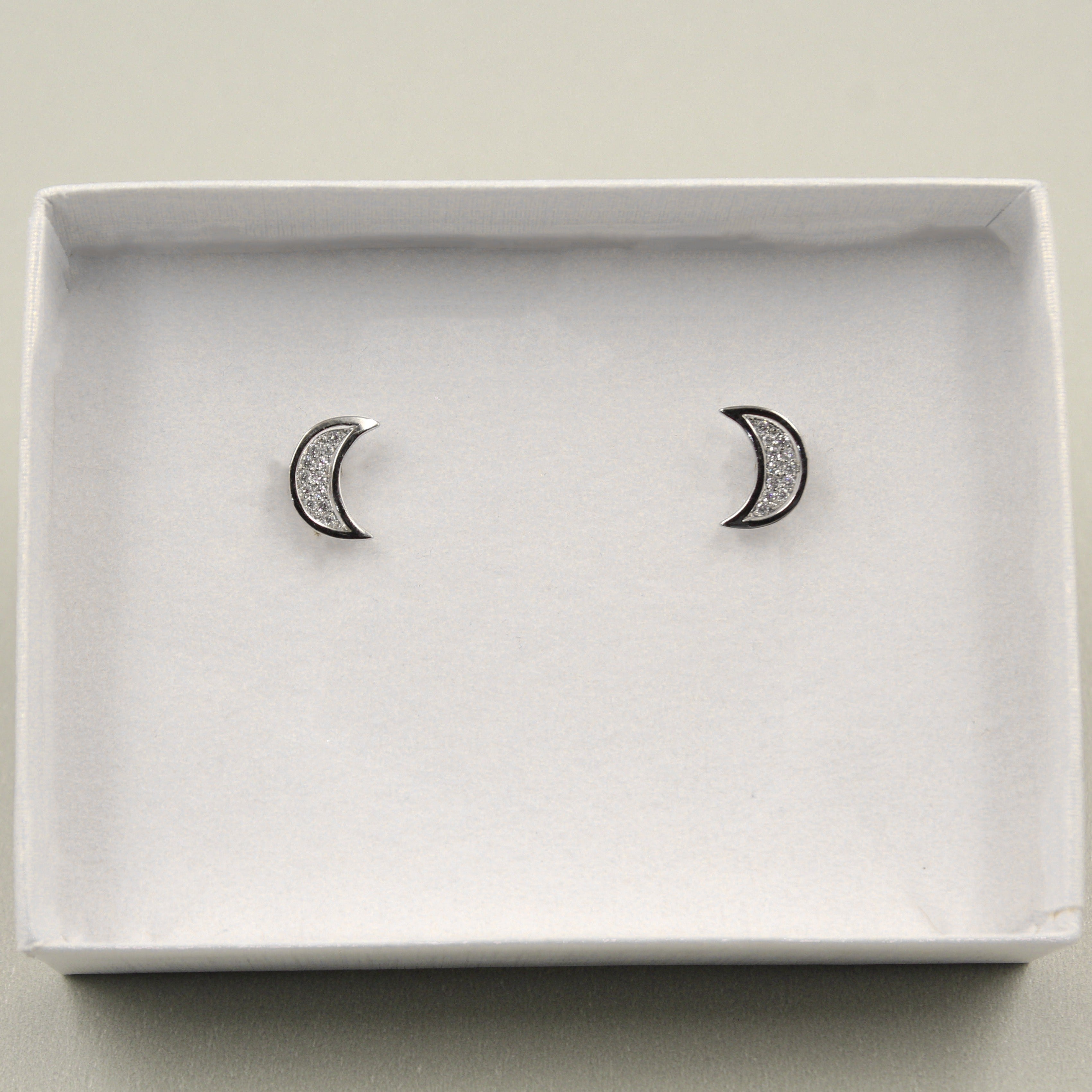 par de aretes de plata con forma de luna creciente, incrustados con pequeños cristales, presentados en una caja blanca con fondo gris.