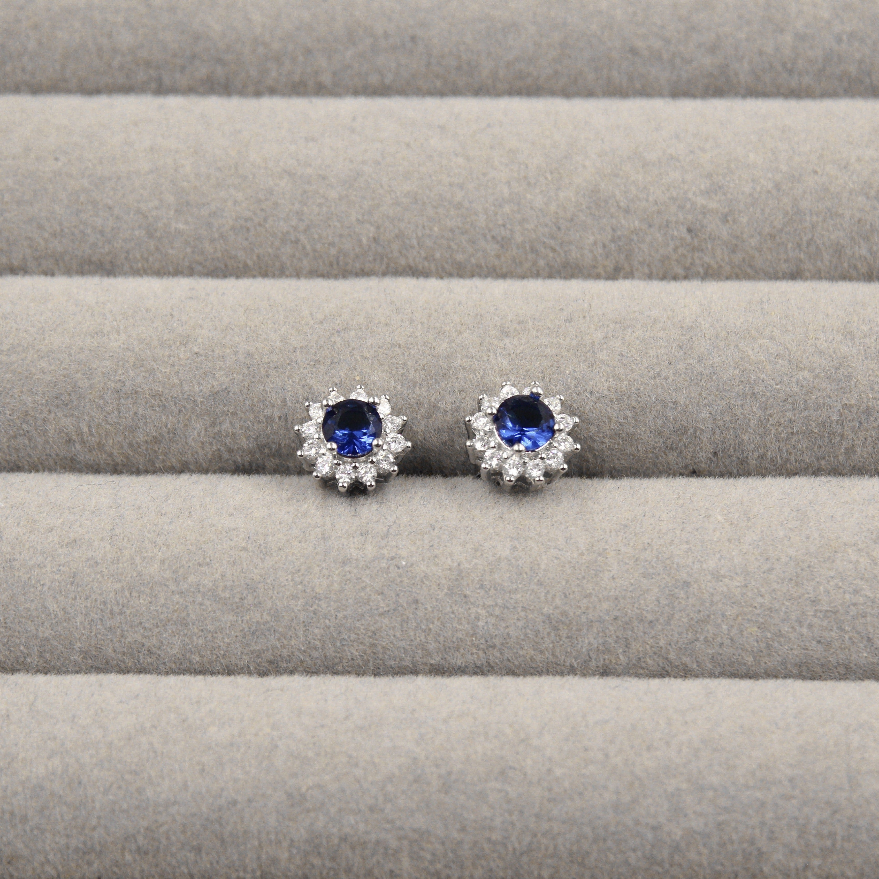 Par de aretes de zafiro azul, rodeados de pequeños diamantes en forma de flor, presentados en un estuche de joyería gris.