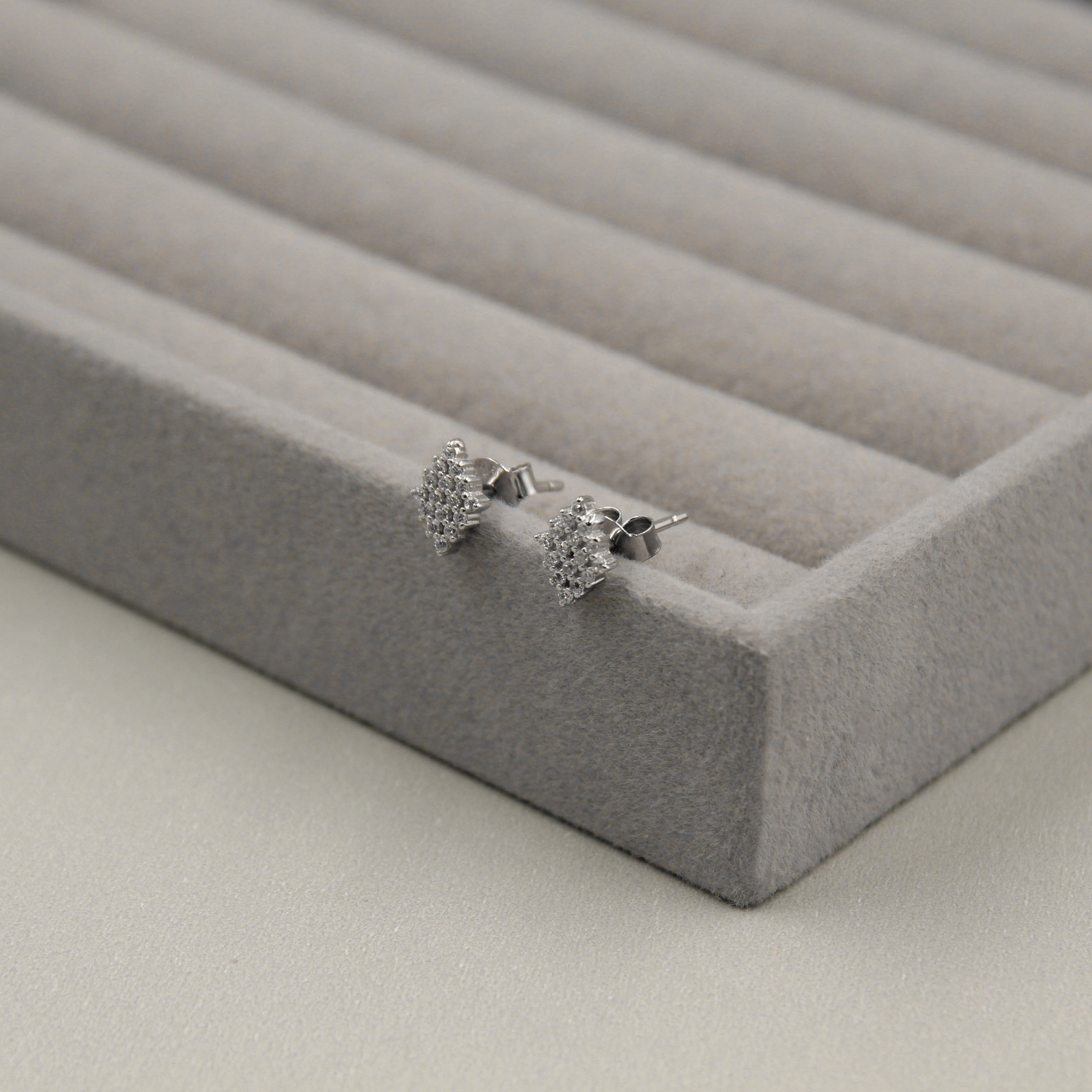Par de aretes pequeños con forma cuadrada, adornados con diamantes, presentados en un estuche de joyería gris en el borde.