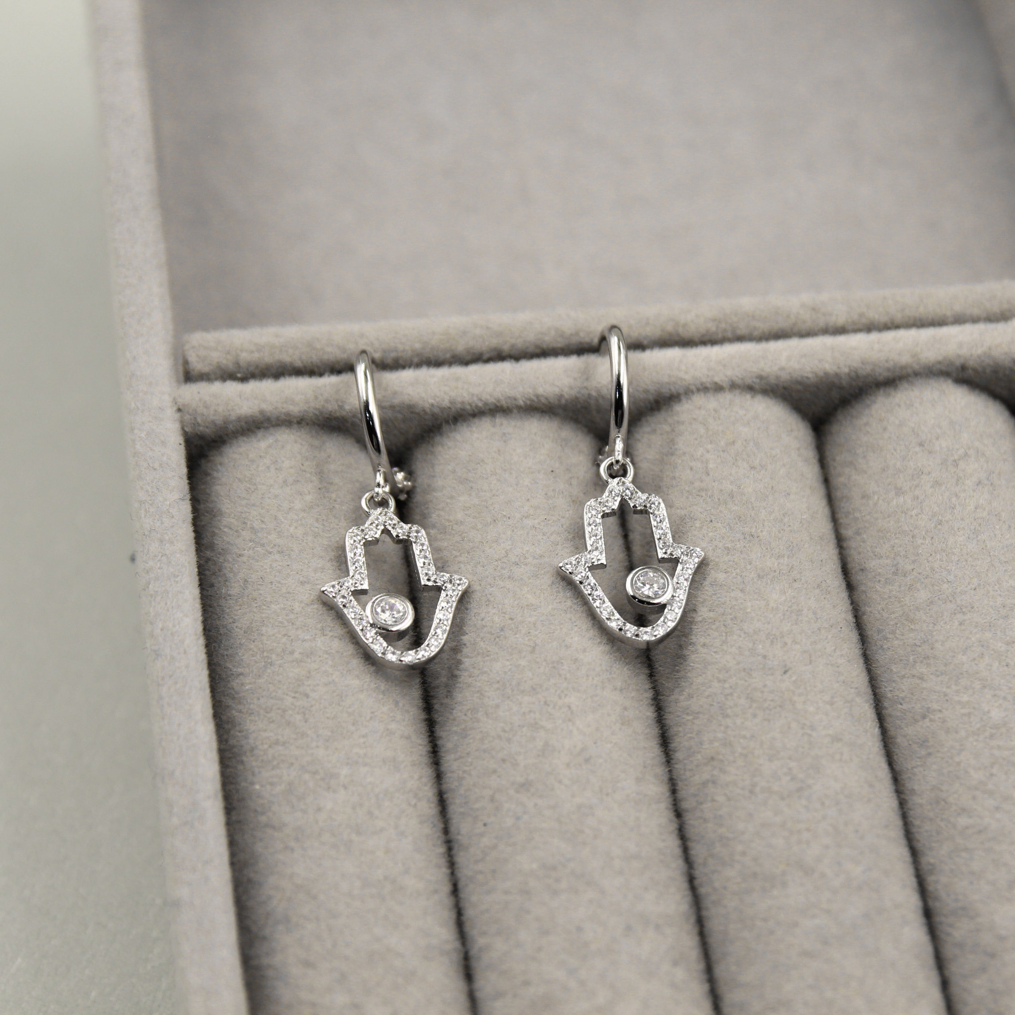 Par de aretes colgantes en forma de mano de Fátima, adornados con pequeños diamantes y un diamante central, presentados en un estuche de joyería gris.