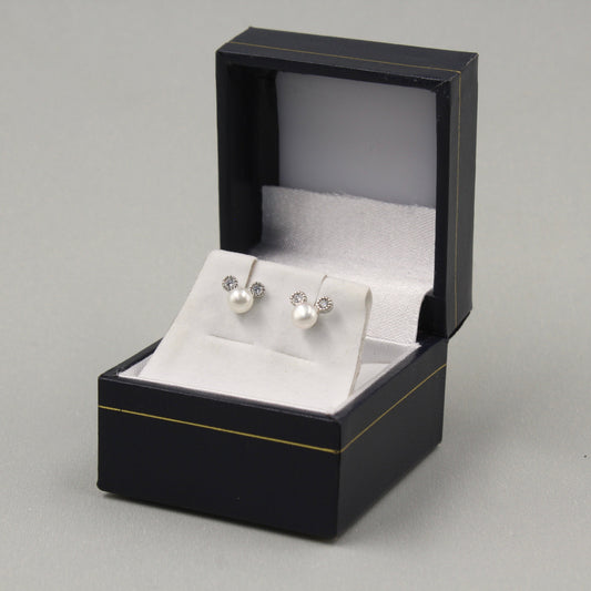 elegante par de aretes de perlas, presentados en una caja de joyería negra con ribetes dorados. Cada arete consiste en una perla blanca con un pequeño adorno de circonia cúbica encima, ofreciendo un diseño clásico y sofisticado.