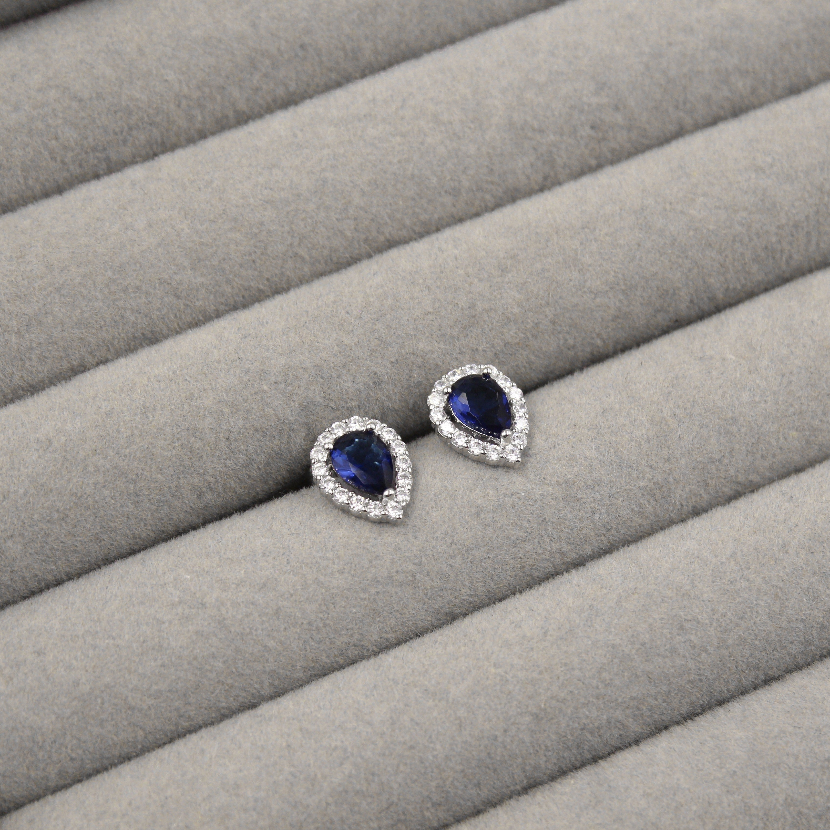 Par de aretes con zafiros azules en forma de lágrima, rodeados de pequeños diamantes, presentados en un estuche de joyería gris.