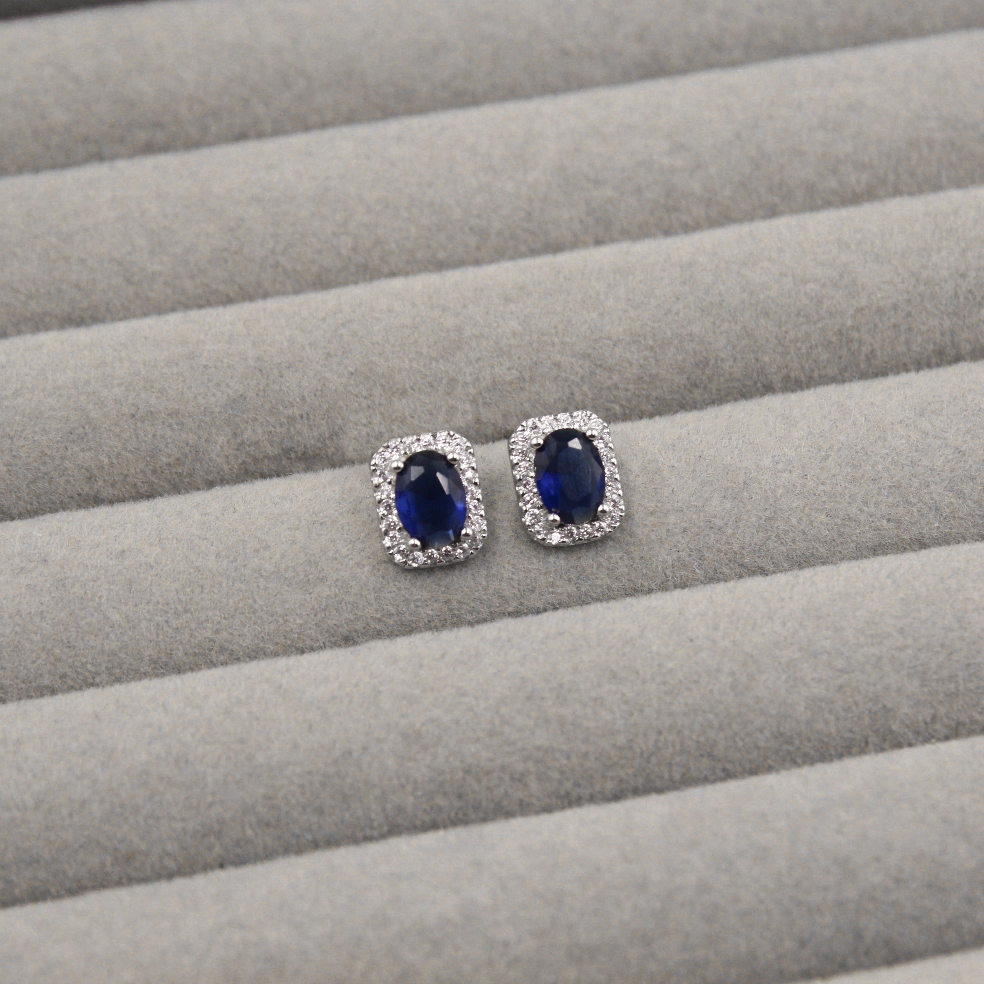 Par de aretes con zafiros azules de corte ovalado, rodeados de pequeños diamantes en un marco rectangular, presentados en un estuche de joyería gris."
