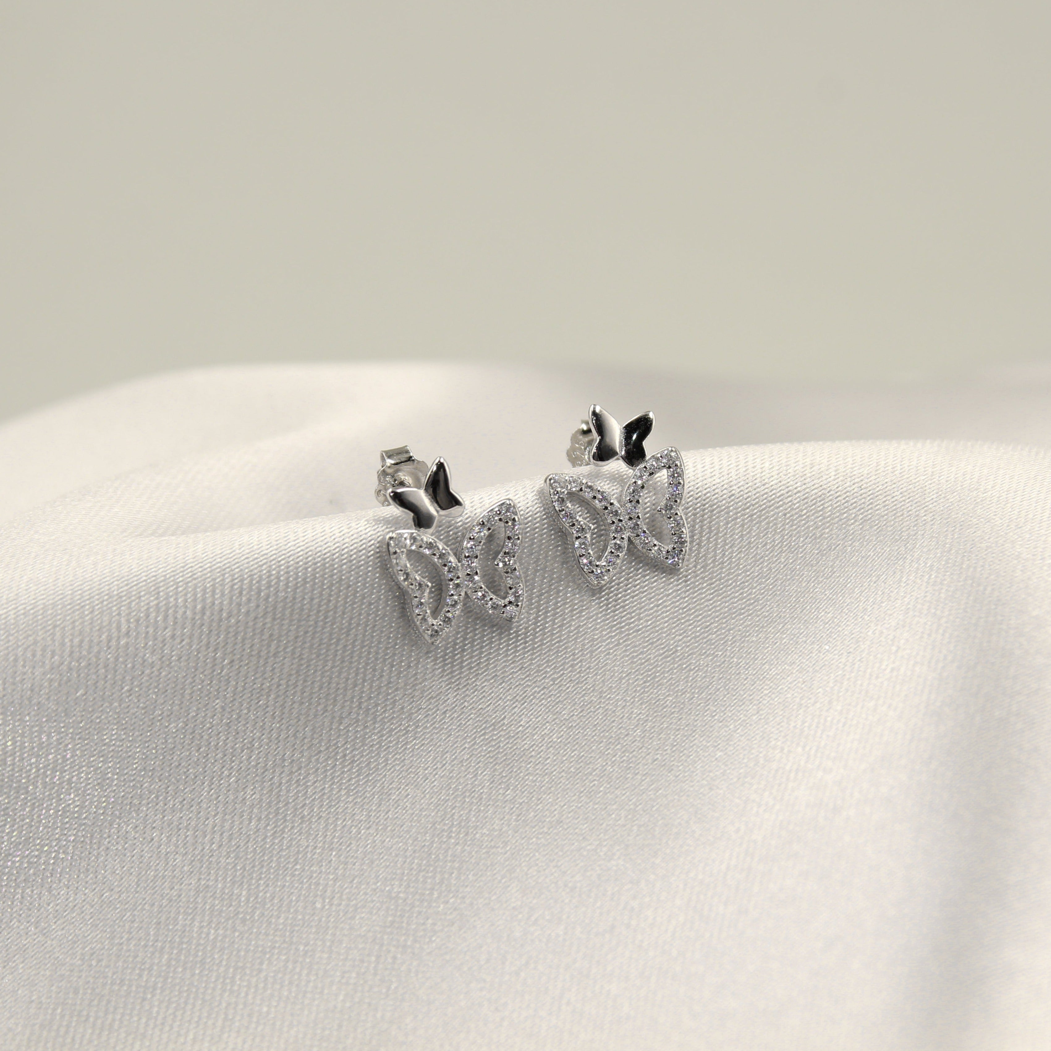 Aretes con diseño de mariposas de dos tonos en plata y negro, destacados contra un fondo de tela blanca.