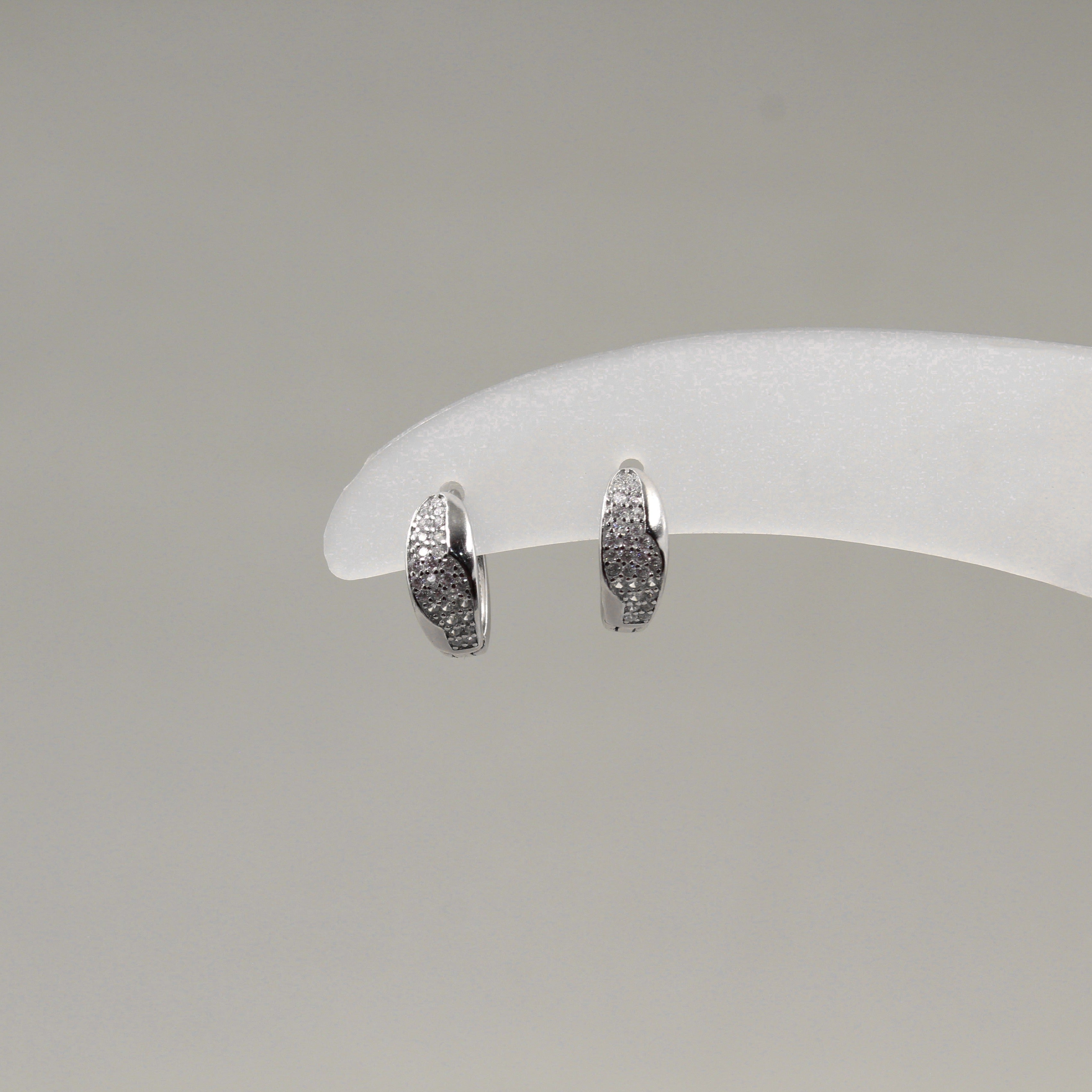 Par de aretes pequeños de aro, adornados con diamantes en un diseño ondulado, exhibidos en un soporte blanco.