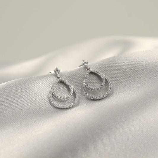 Aretes colgantes en forma de gota doble con incrustaciones de diamantes sobre un fondo de tela blanca.